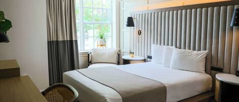A hotel room with a large bed, a desk, a chair, a small table with a plant, and a window with curtains.