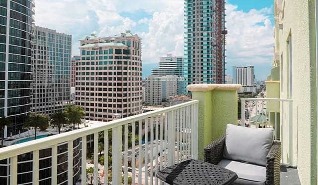 A balcony with a view of high-rise buildings, a white railing, and a wicker chair with cushions.