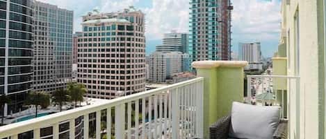A balcony with a view of high-rise buildings, a white railing, and a wicker chair with cushions.