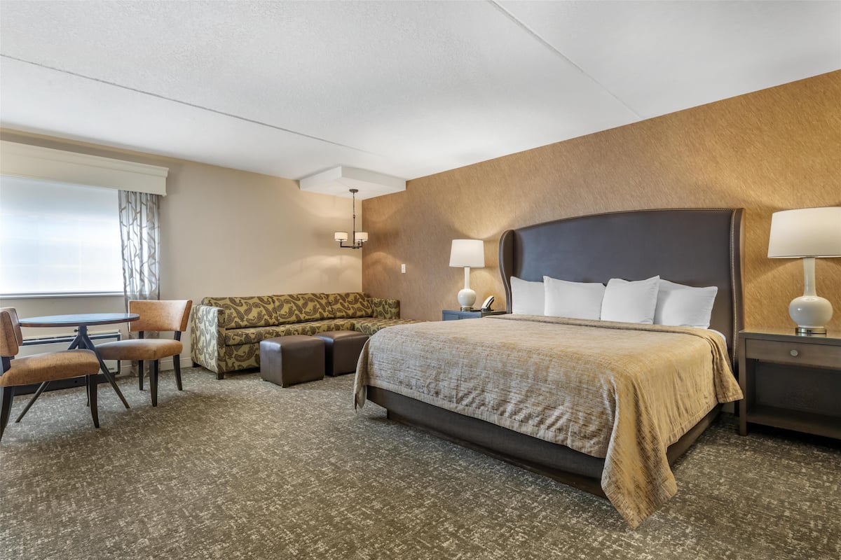 A hotel room with a large bed, a sofa, a dining table with chairs, and a small table with a lamp.