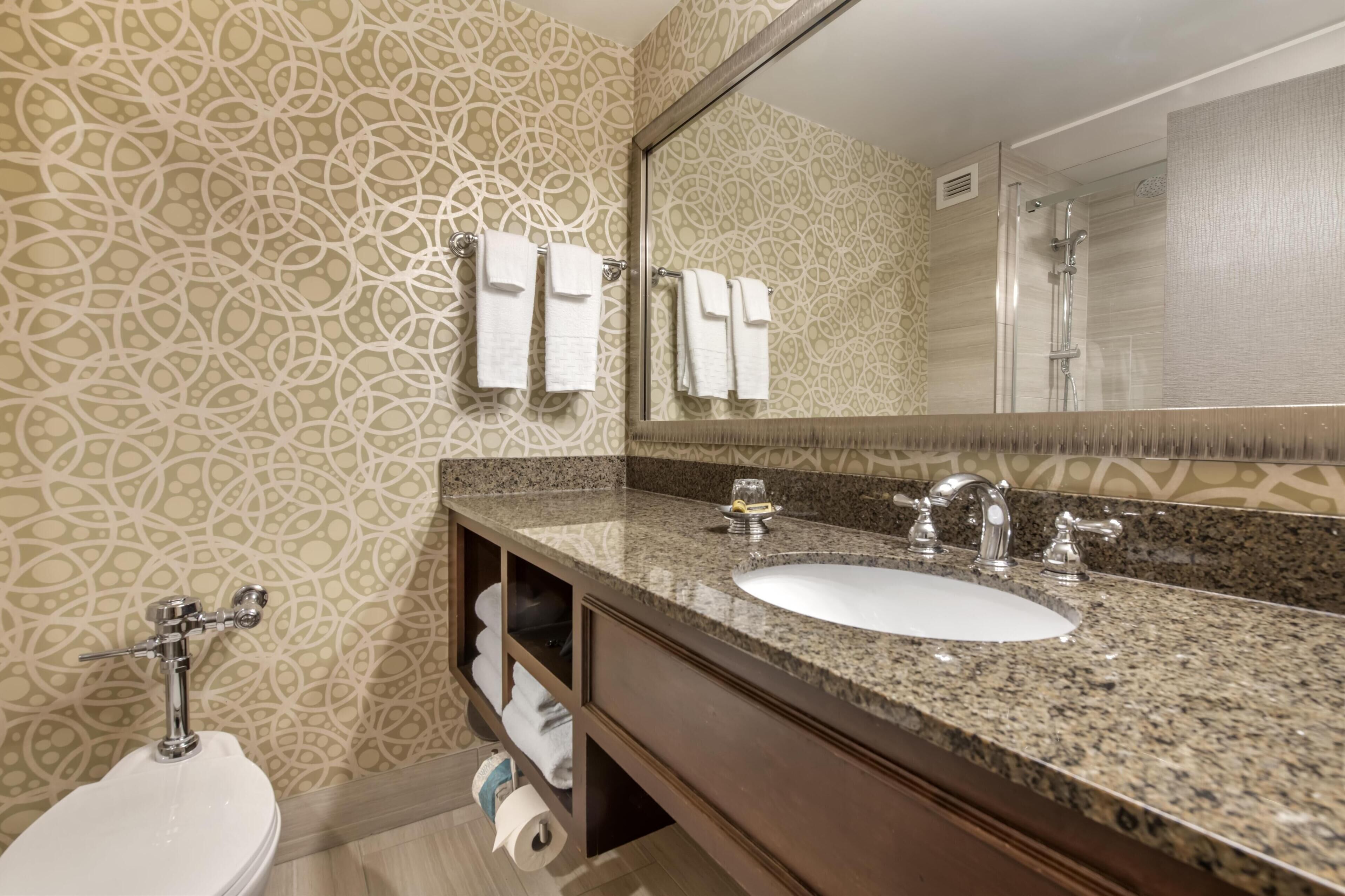 A bathroom with a patterned wall, a sink with a mirror, a toilet, and a shower.