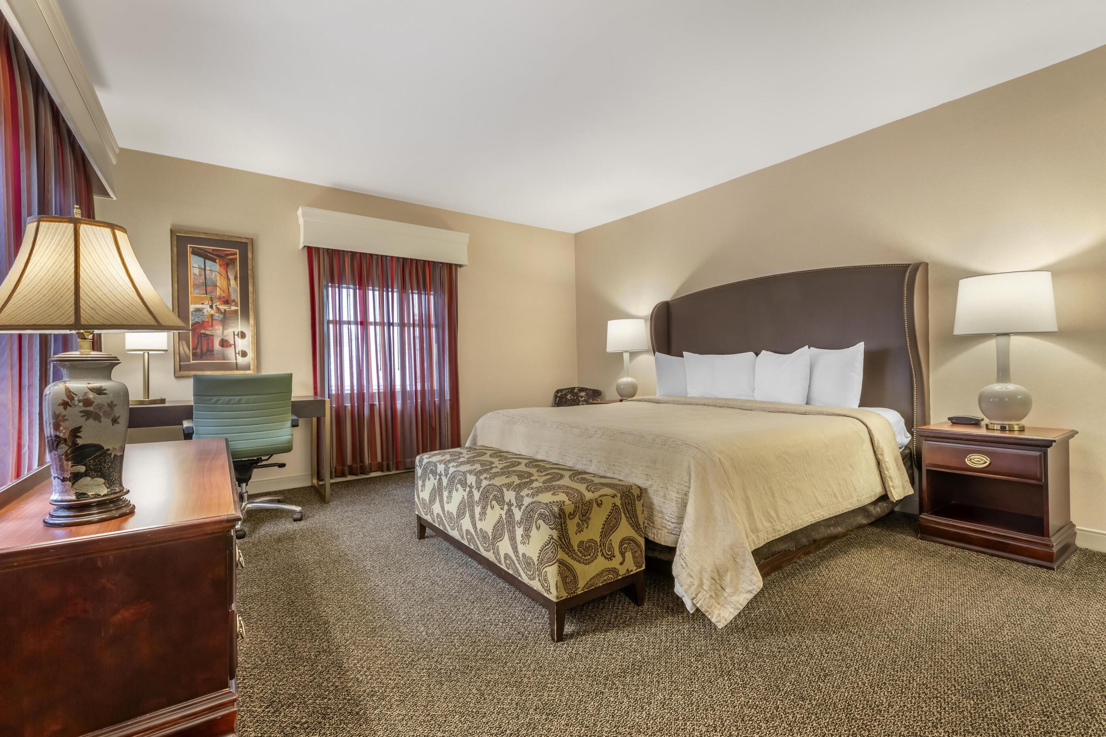 Habitación de hotel con una cama grande, un escritorio, una silla, una mesita de noche con lámparas y una ventana con cortinas.