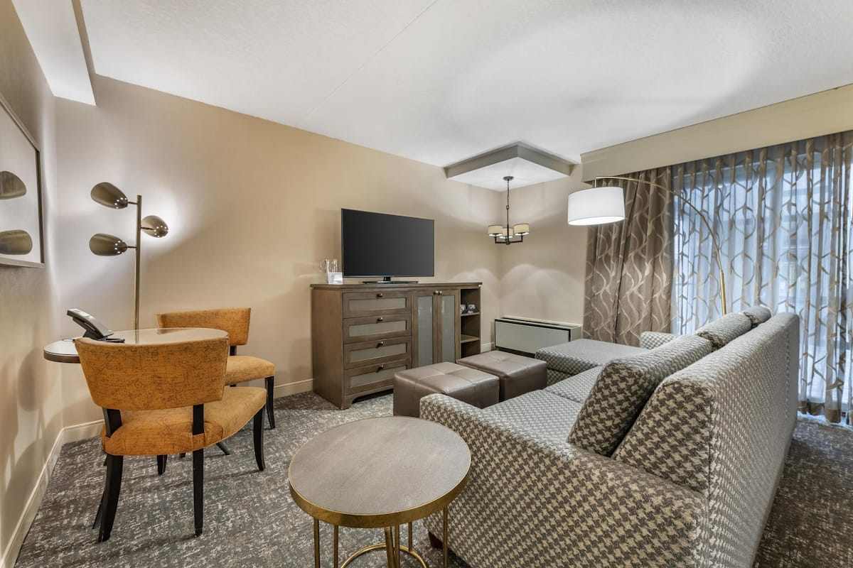 A hotel room with a sofa, two chairs, a small round table, a TV, a cabinet, and a window with curtains.