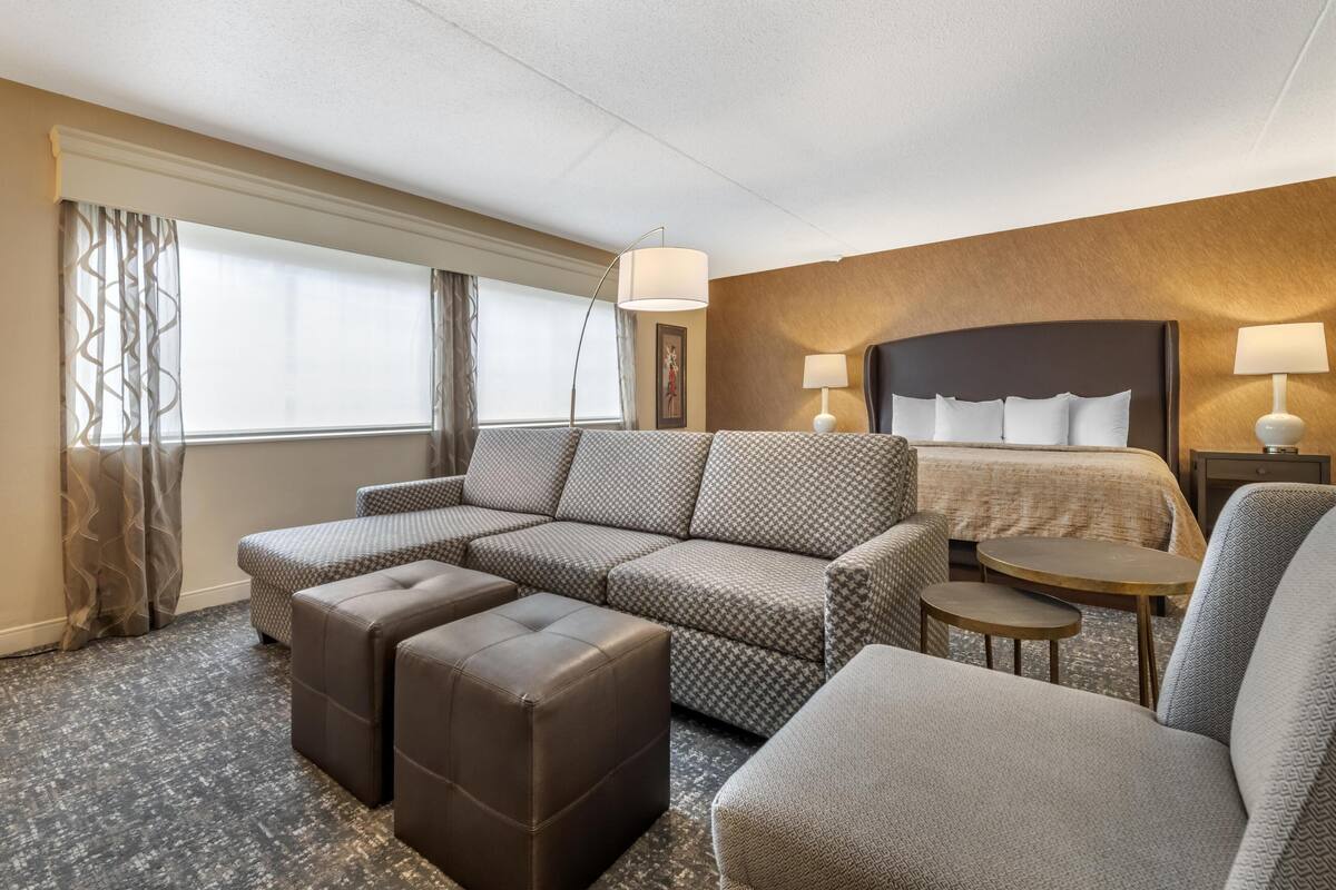 A hotel room with a large sectional sofa, a bed with a headboard, two bedside tables with lamps, and a view of the city through large windows.