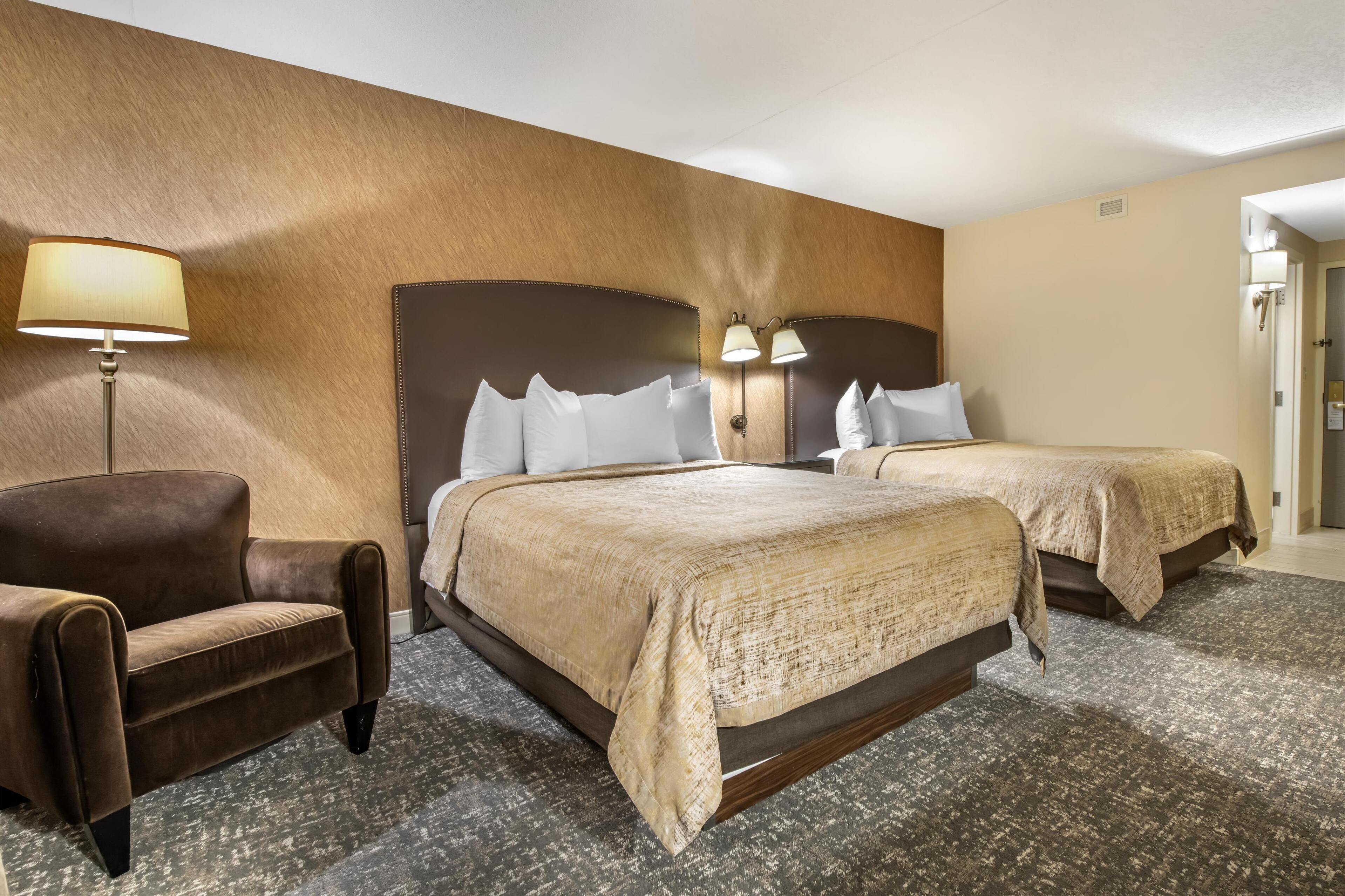 A hotel room with a large bed, two bedside tables, a brown armchair, and a lamp.