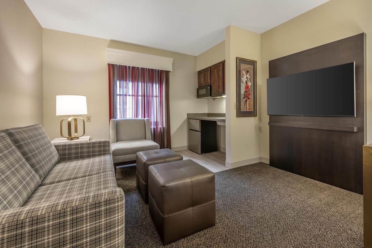 A hotel room with a sofa, ottoman, kitchenette, flat-screen TV, and a painting on the wall.