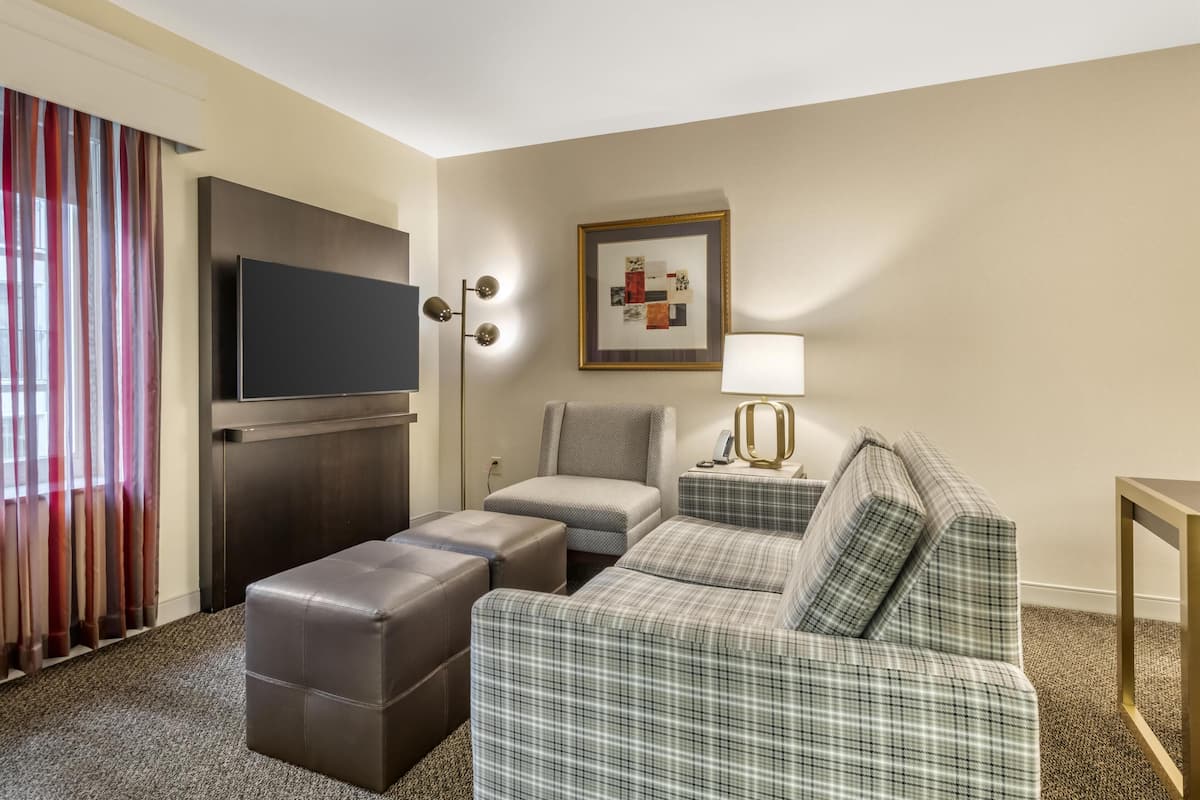 A hotel room with a flat-screen TV, a sofa, an ottoman, a lamp, and a framed artwork on the wall.