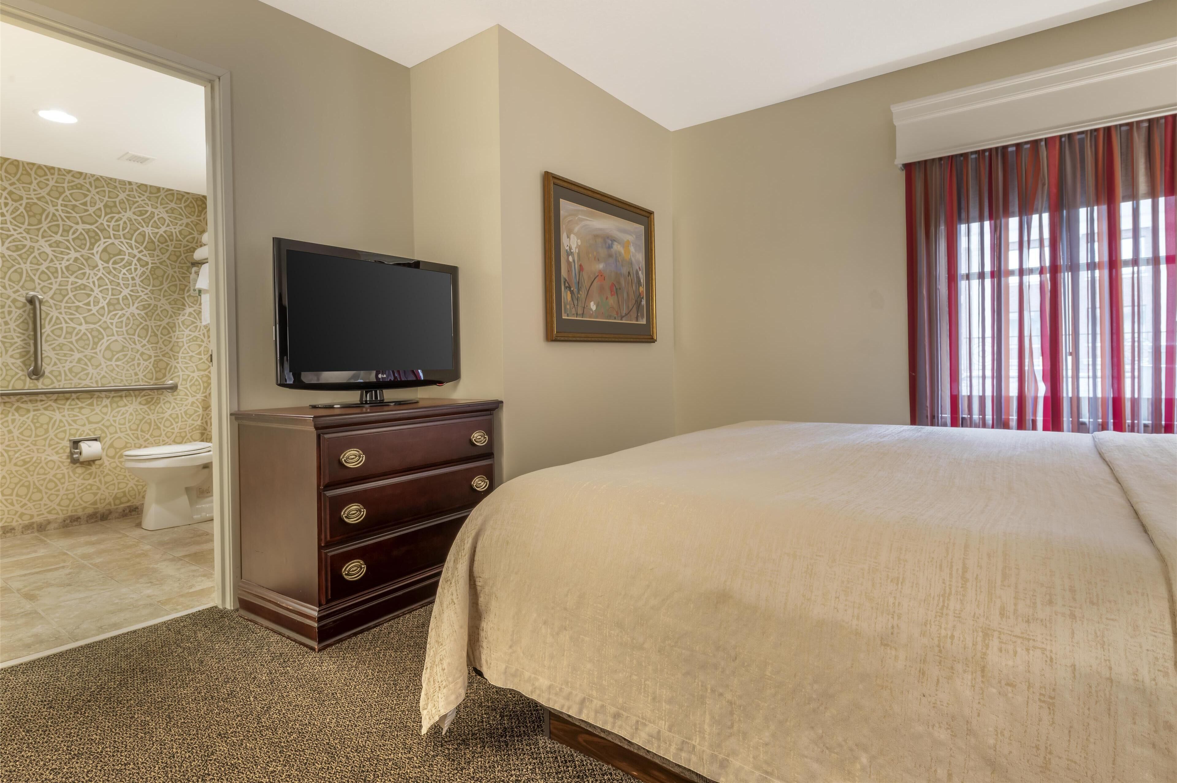 Habitación de hotel con una cama grande, un aparador con cajones, un televisor sobre el aparador, un cuadro en la pared y una ventana con cortinas.