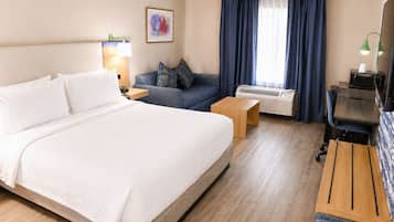 A hotel room with a large bed, a sofa, a desk, and an air conditioning unit.