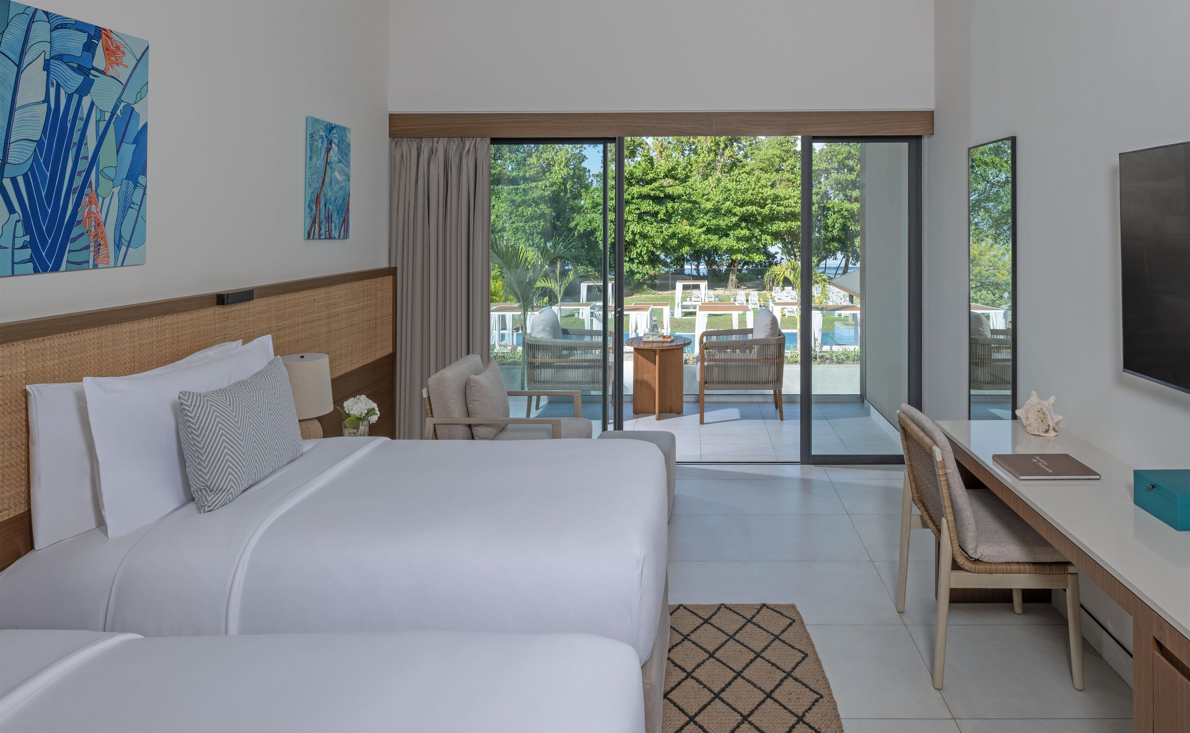 A hotel room with a bed, a desk, a chair, and a view of an outdoor area with a pool and lounge chairs.