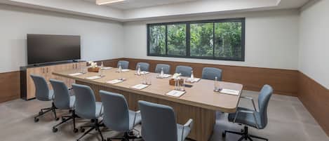 A conference room with a long table, chairs, a TV, and a window with a view of greenery.