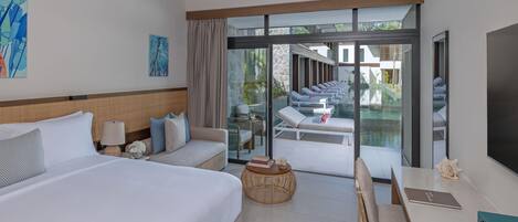 A hotel room with a bed, a sofa, a dining table, a TV, and a view of a pool and outdoor seating.