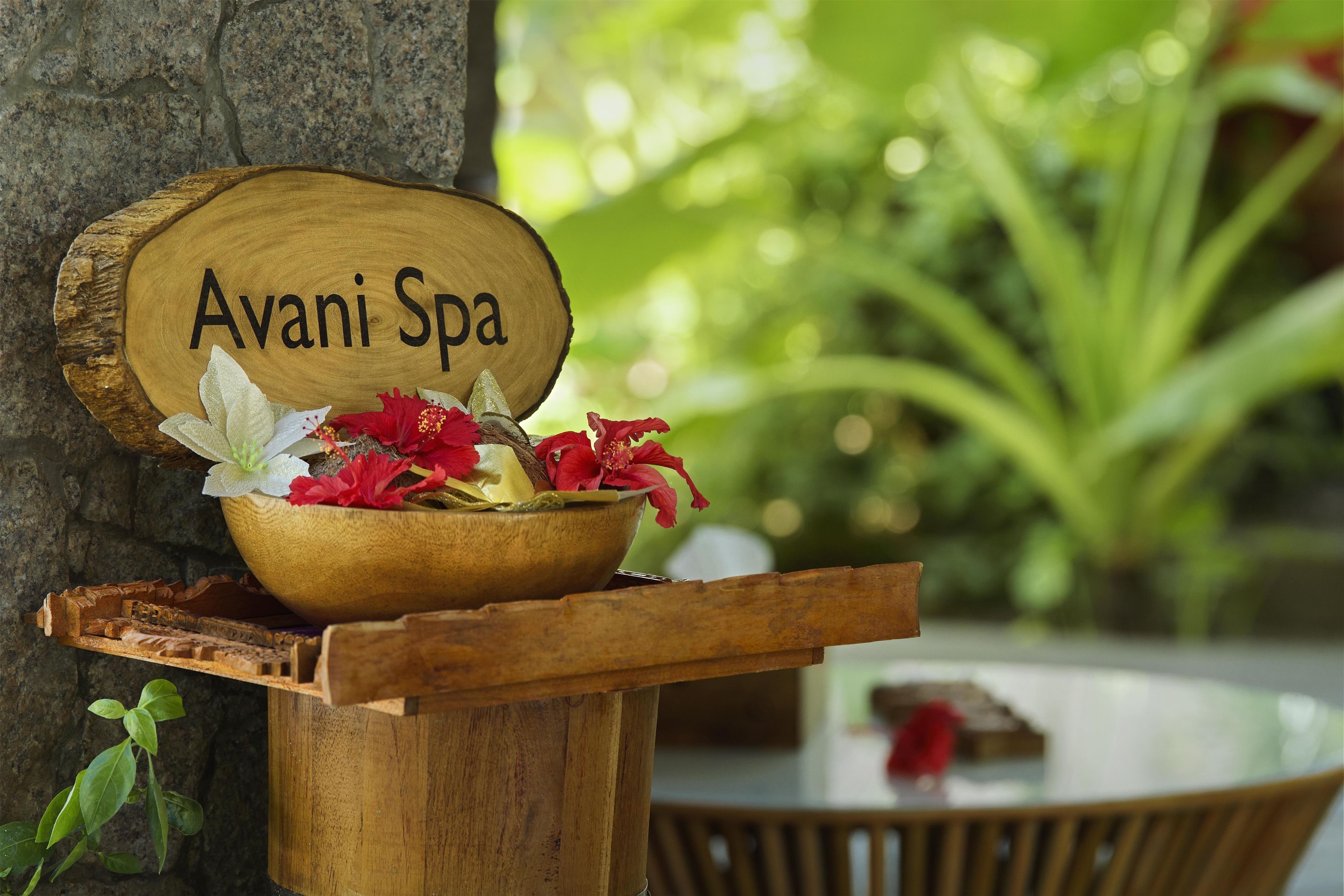 Un letrero de madera con la inscripción 'Avani Spa', colocado sobre un atril de madera con un cuenco que contiene flores rojas y una flor blanca.