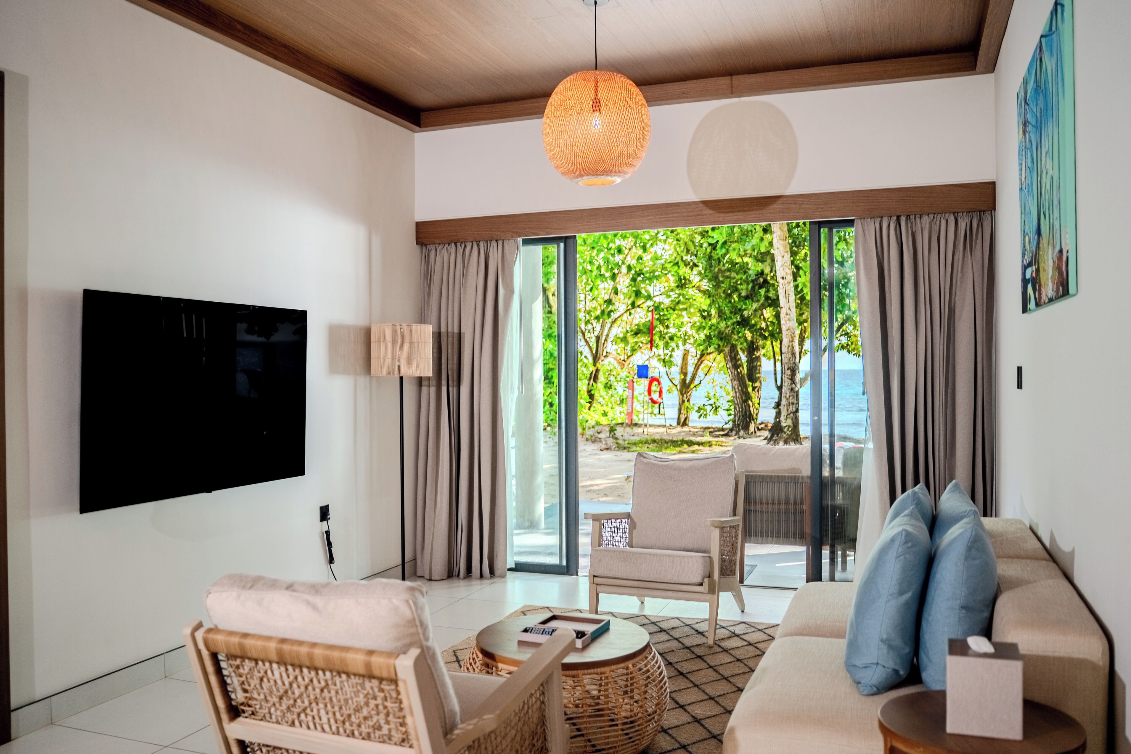 A modern living room with a flat-screen TV, a sofa, a chair, and a table. There is a large window with curtains, and a view of a beach and trees outside.