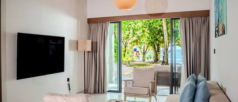 A modern living room with a flat-screen TV, a sofa, a chair, and a table. There is a large window with curtains, and a view of a beach and trees outside.