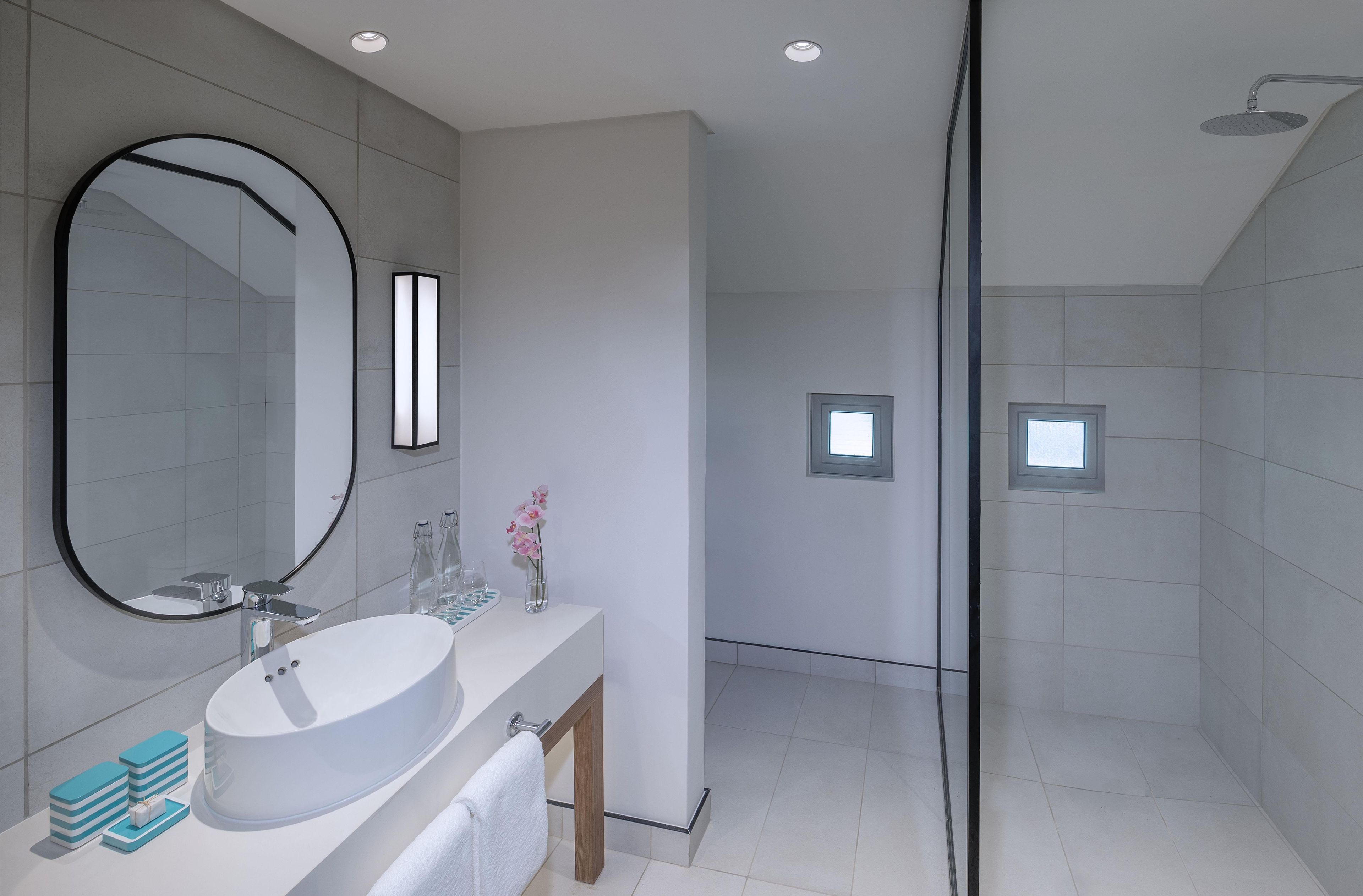 Un baño moderno con un espejo redondo grande, un lavamanos blanco y una zona de ducha con mampara de cristal.
