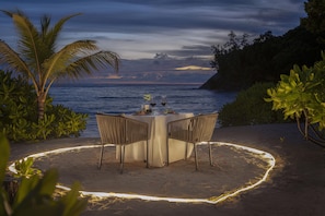 A table set for two with chairs, a candle, and a glass of wine on a sandy beach with a view of the ocean and palm trees. - Avani Plus Barbarons Seychelles Resort (Mahé Island)