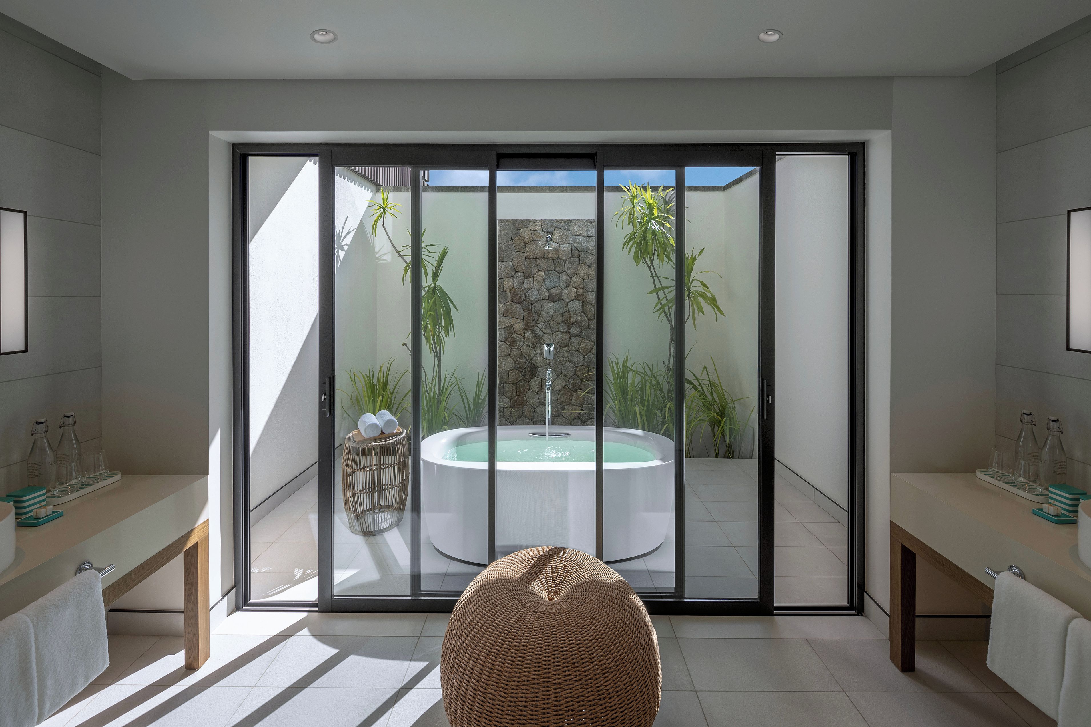 Un baño moderno con un amplio cerramiento de vidrio corredera que da a un área de spa al aire libre, una bañera de hidromasaje integrada y un banco de madera.