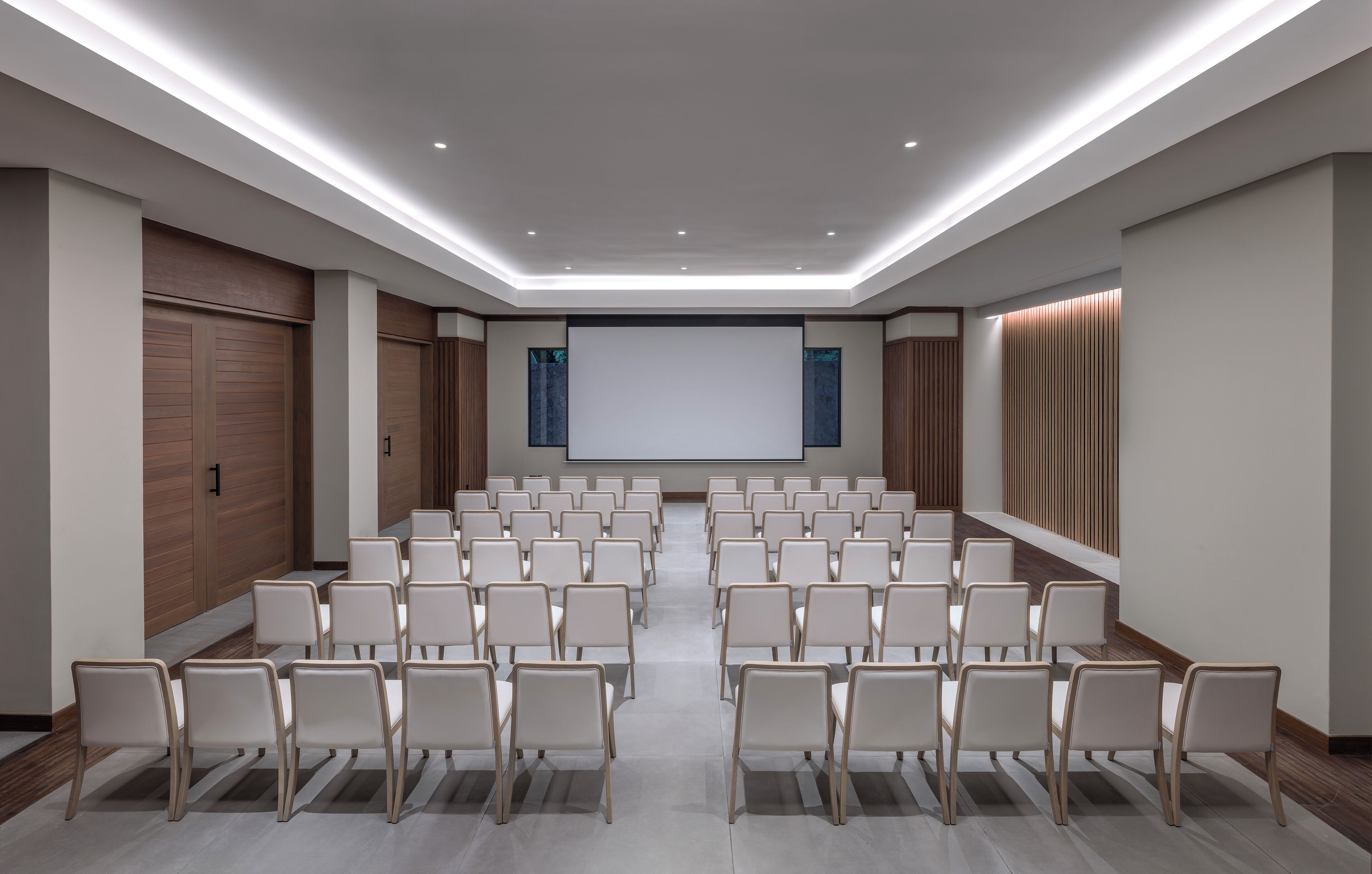 A conference room with rows of chairs facing a large screen, wooden panel walls, and recessed ceiling lighting.