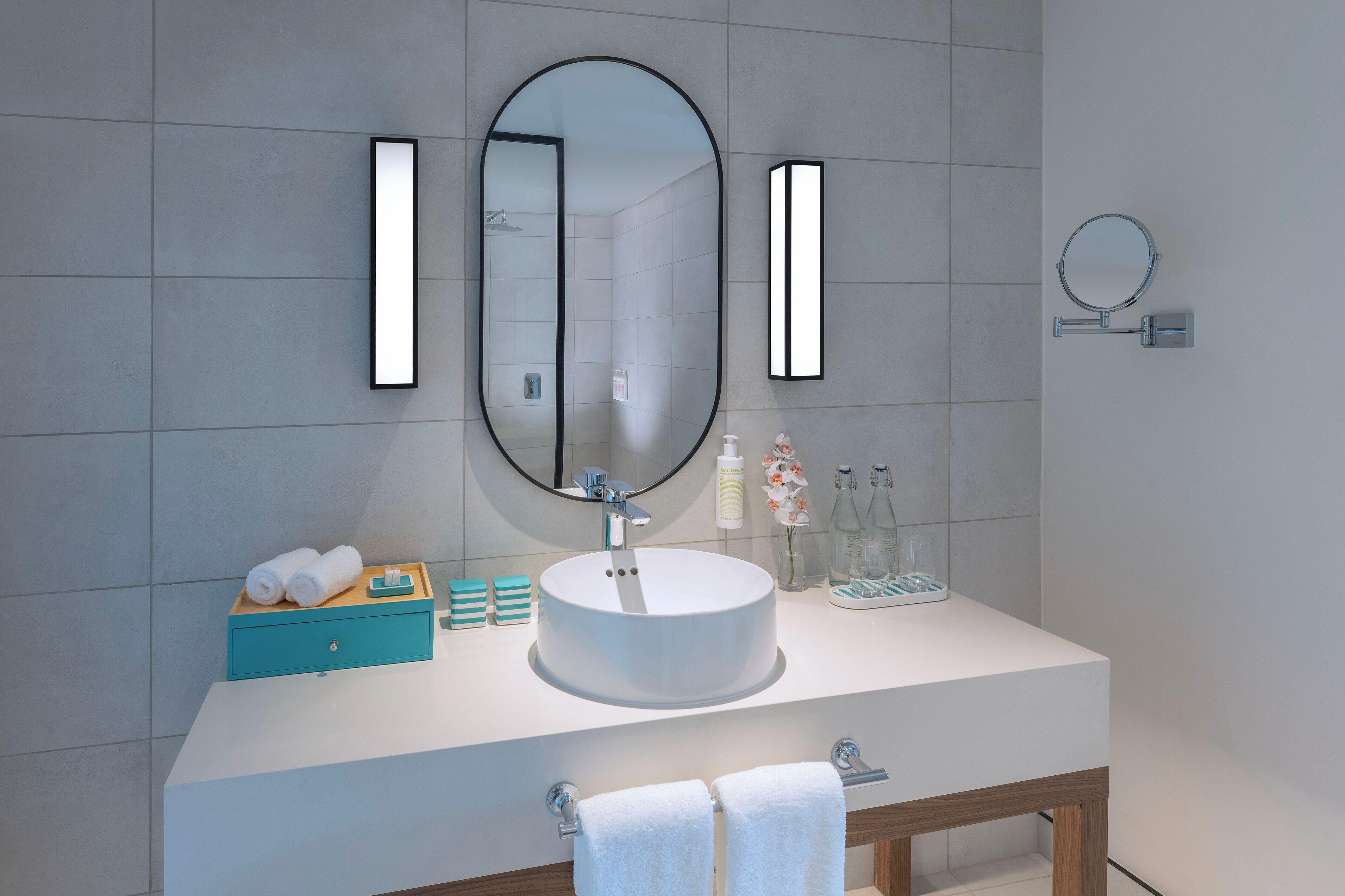A bathroom with a large oval mirror, a rectangular mirror, and a small round mirror. There is a white sink with a wooden vanity, a towel rack, and a wooden stool.