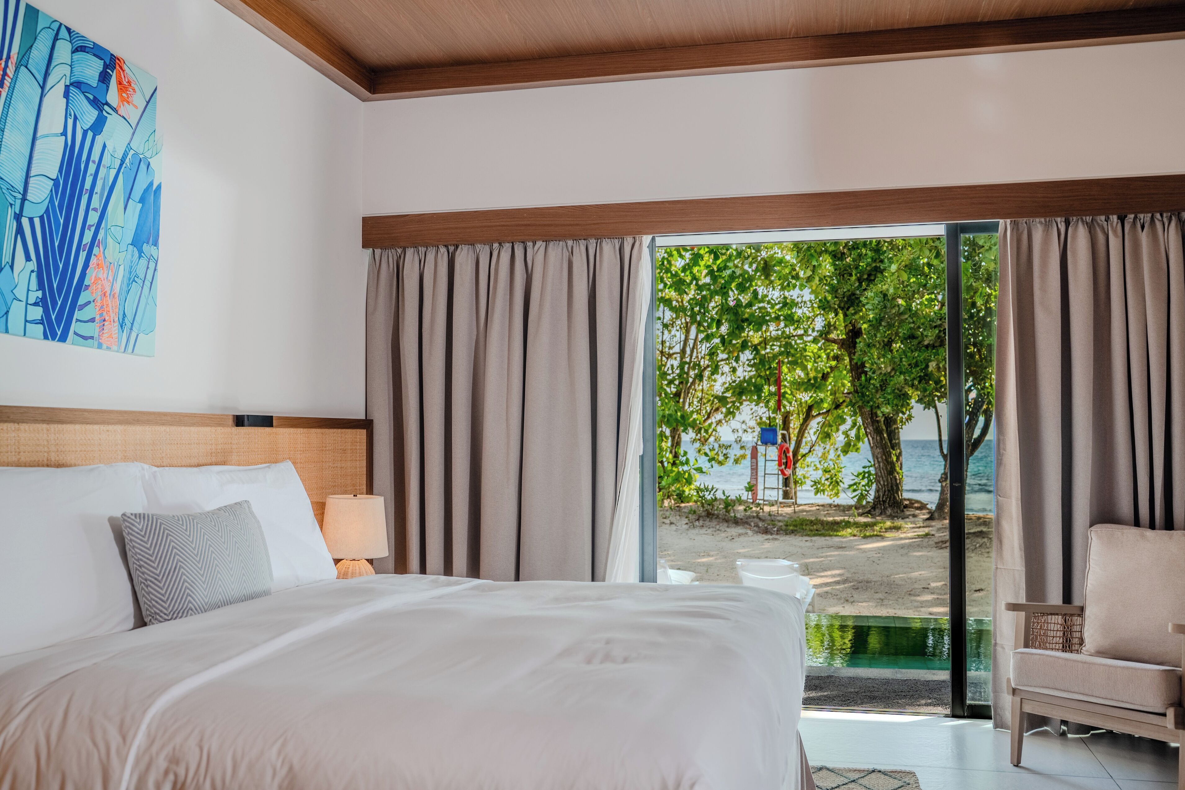 Ett hotellrum med en stor säng, en stol, en lampa och utsikt över en strand genom fönstret.