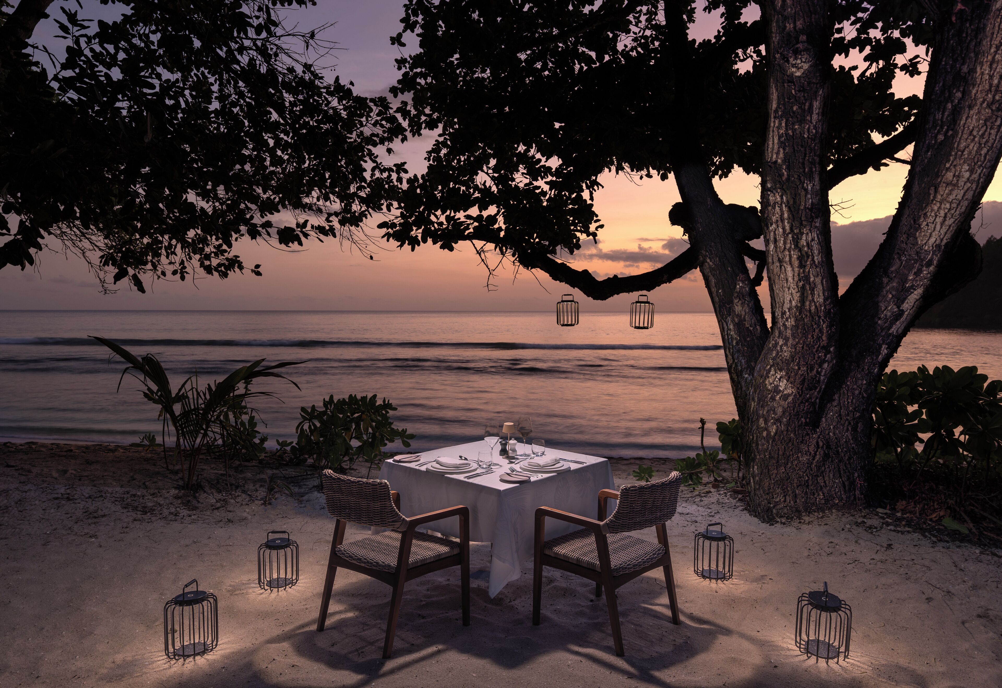 A table set for two under a tree with lanterns, overlooking a beach at sunset.