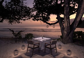 Mesa para dos bajo un árbol con faroles, con vistas a una playa al atardecer.