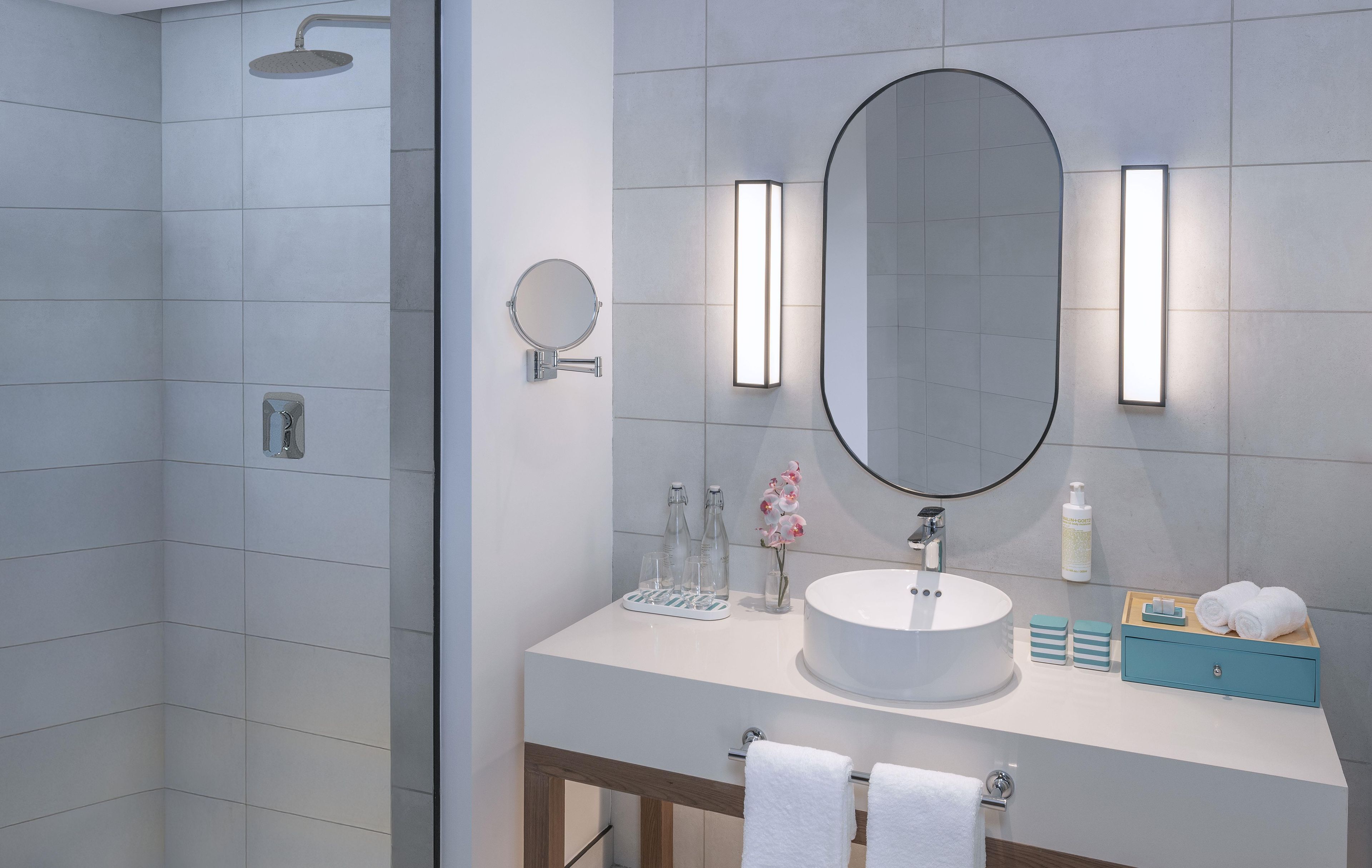 Un baño moderno con ducha, un espejo redondo, un espejo ovalado grande y un lavabo blanco con tocador de madera.