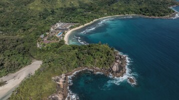 Vista aérea de un complejo turístico frente al mar, con playa, piscina y abundante vegetación circundante.