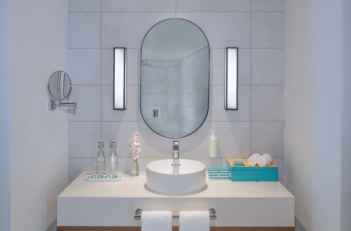 Un baño con un espejo redondo, dos luces empotradas, un lavamanos con encimera blanca y un caja azul para guardar objetos.