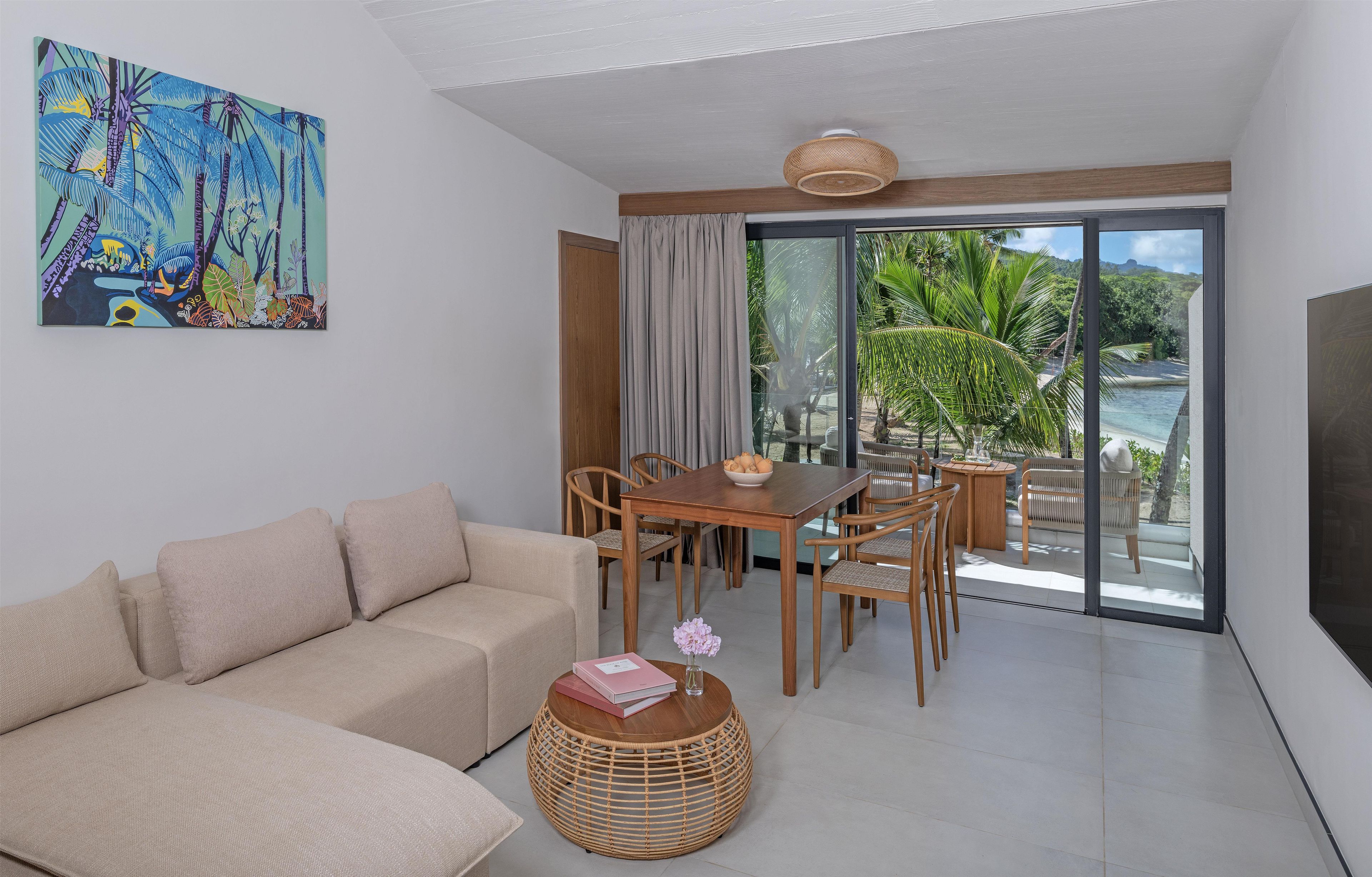 A modern living room with a sofa, dining table, chairs, and a view of a tropical landscape through large glass doors.