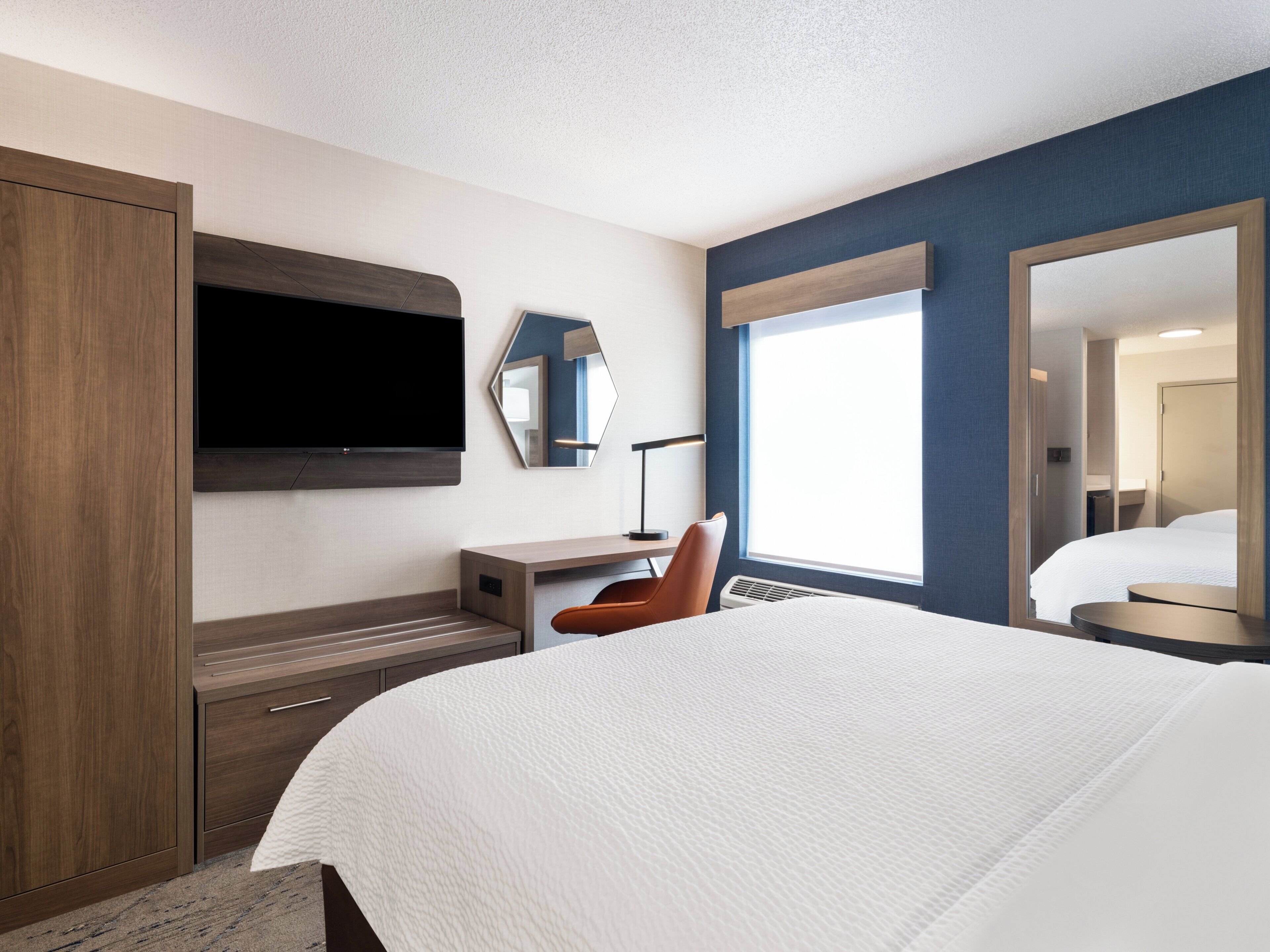 Une chambre d’hôtel dotée d’un grand lit, d’un téléviseur à écran plat fixé au mur, d’un bureau avec une chaise et d’une fenêtre laissant entrer la lumière naturelle.
