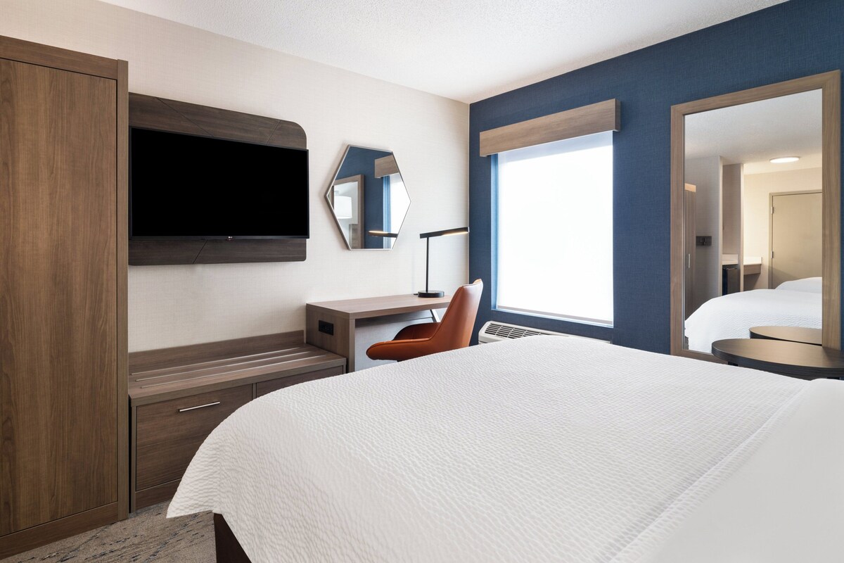 A hotel room with a large bed, a TV mounted on the wall, a desk with a chair, and a large mirror.