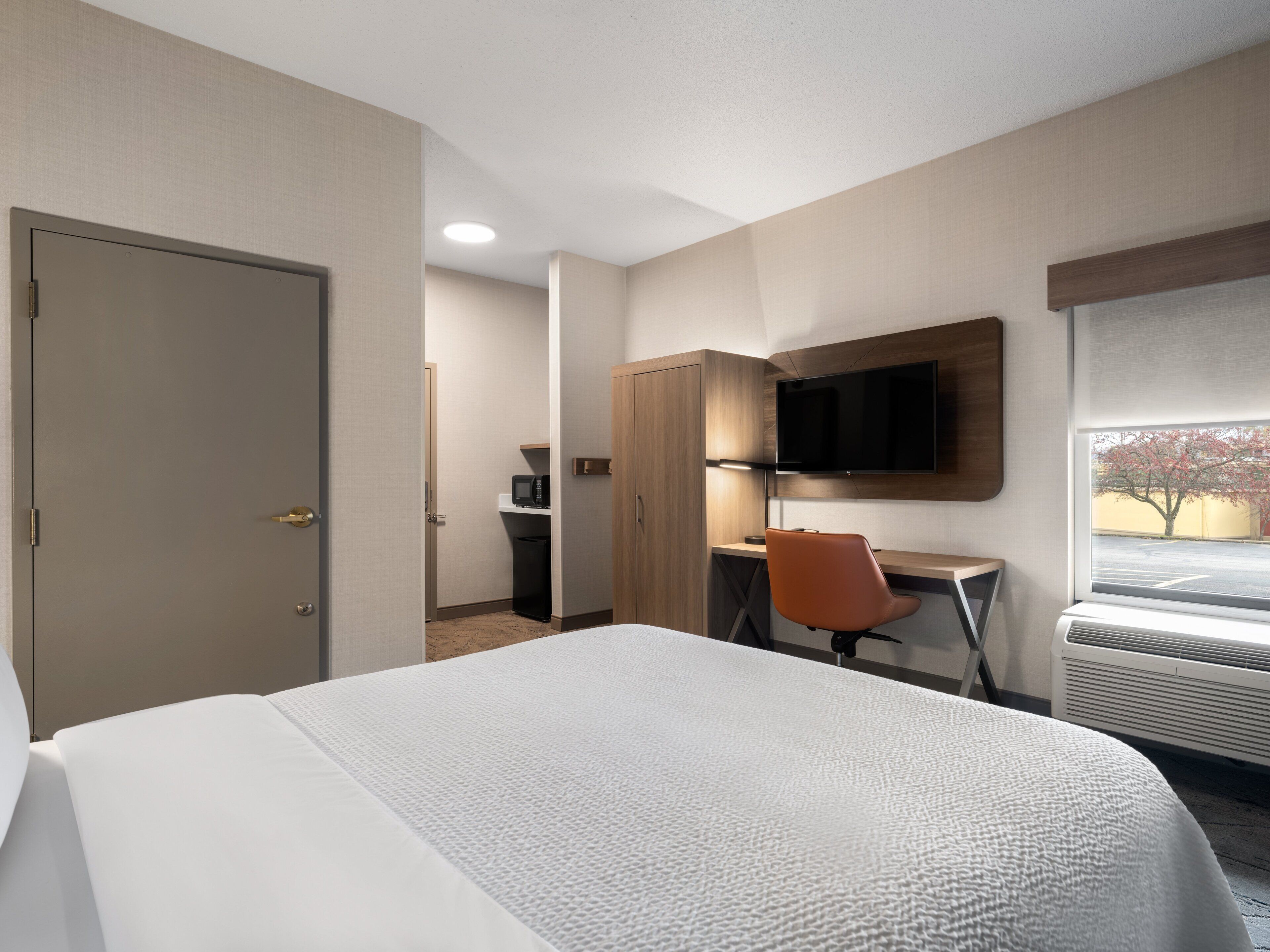 A hotel room with a bed, a desk, a chair, a television, and a window with a view of a parking lot.