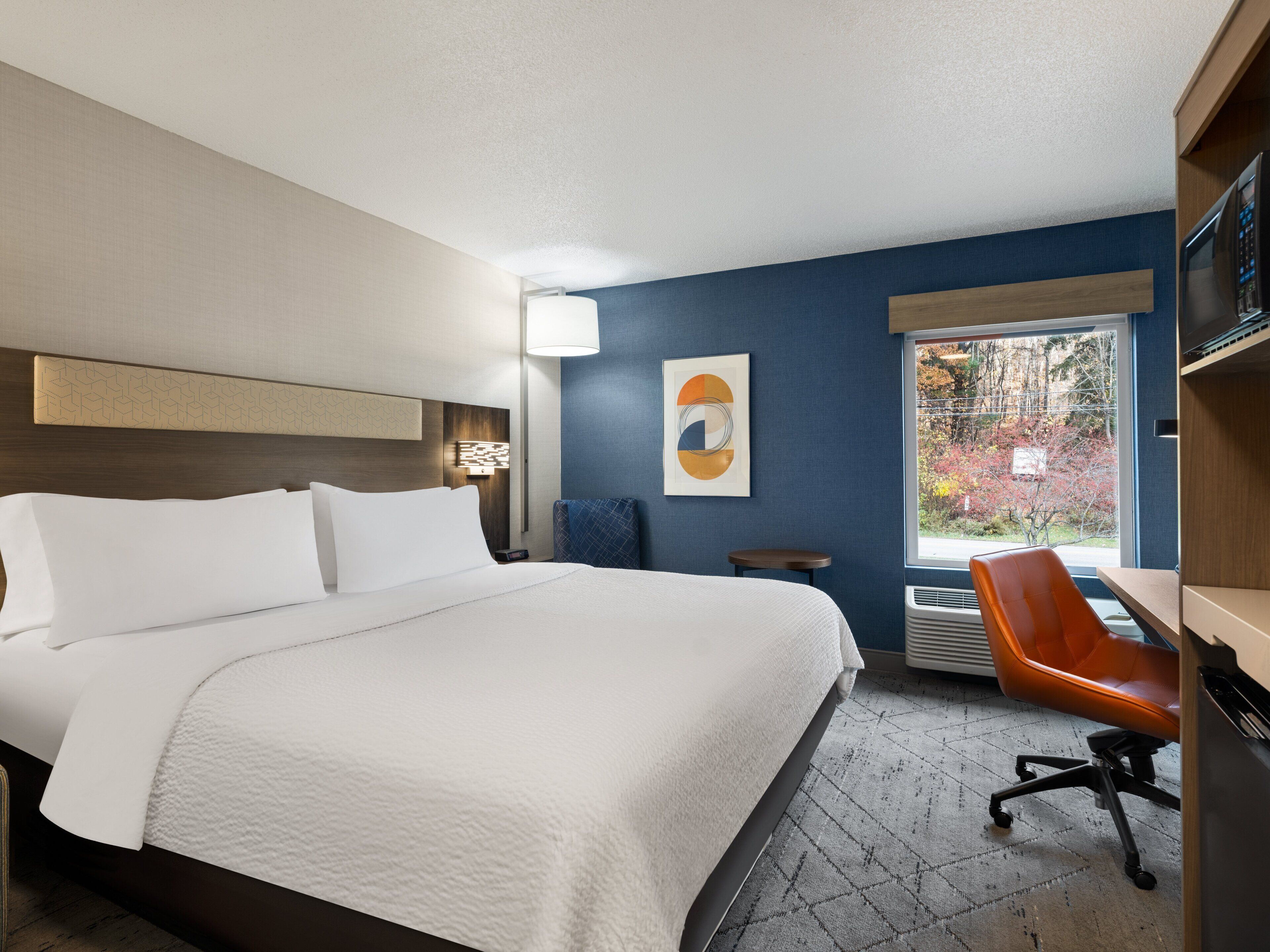 Quarto de hotel com uma cama grande, uma escrivaninha com cadeira, uma janela com vista para árvores e uma obra de arte na parede.