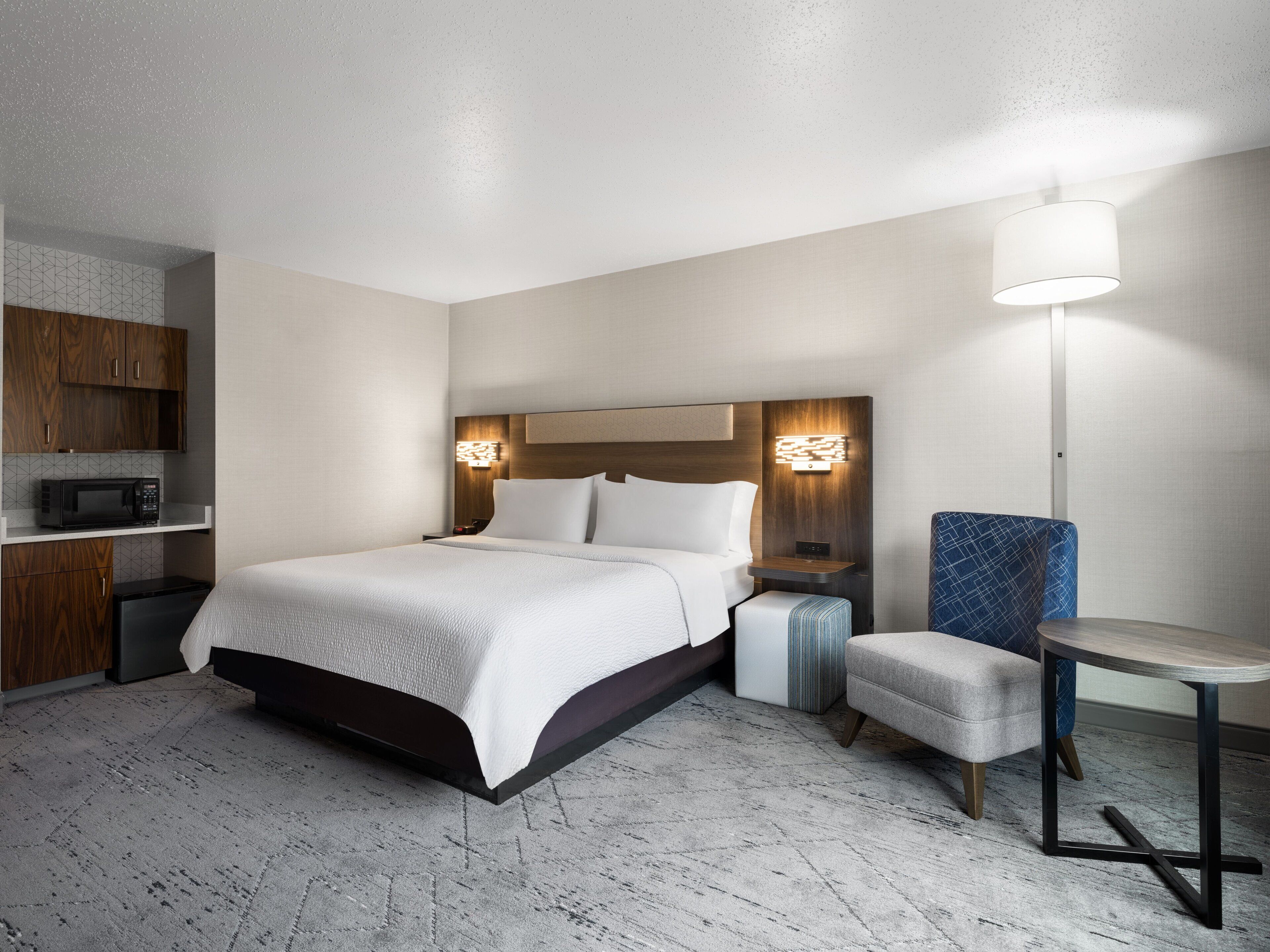 A hotel room with a bed, bedside table, lamp, and a kitchenette area.