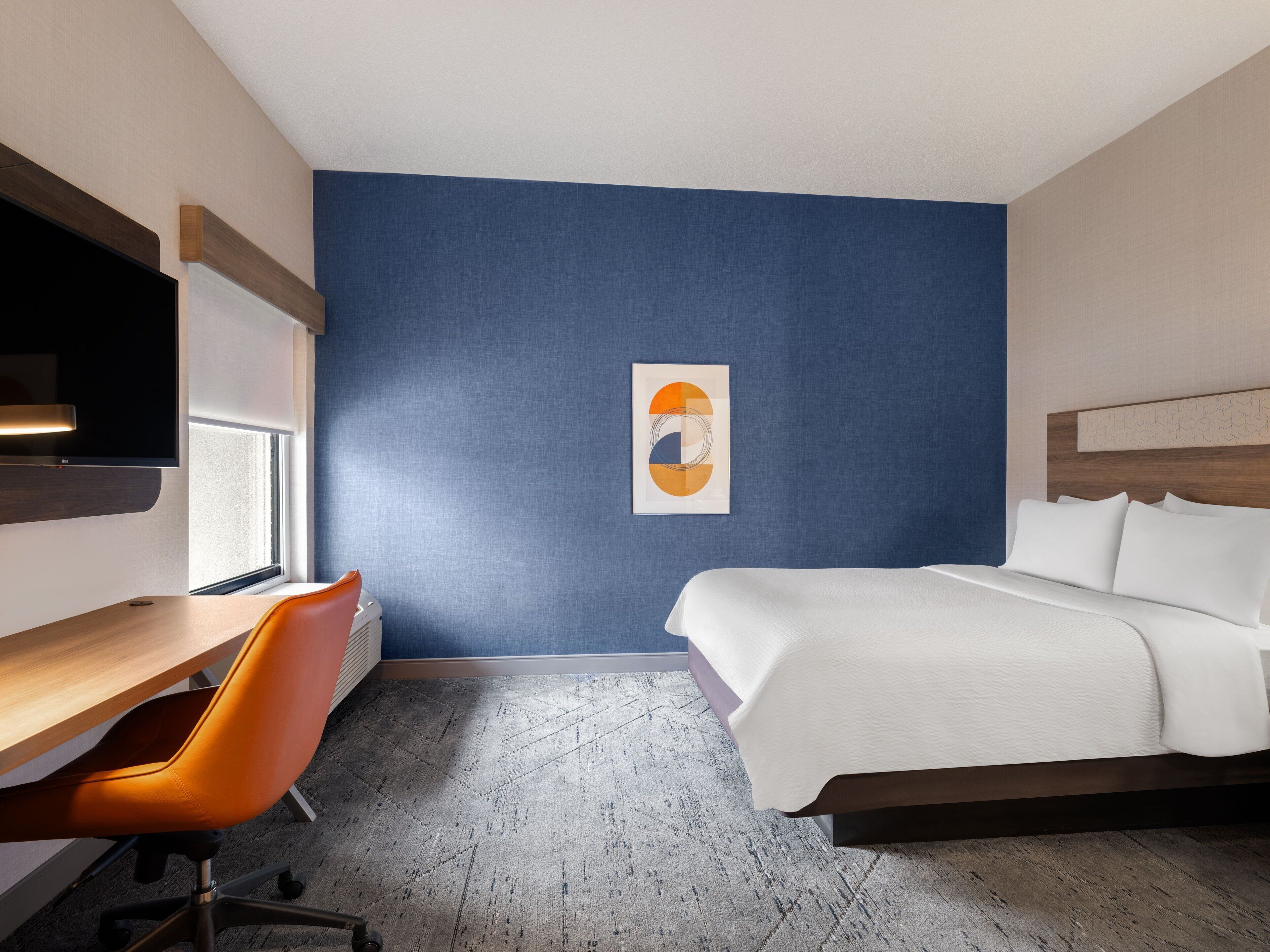 Ein Hotelzimmer mit einem Bett, einem Schreibtisch, einem orangefarbenen Sessel, einem Fernseher und einer Wand in Blautönen.