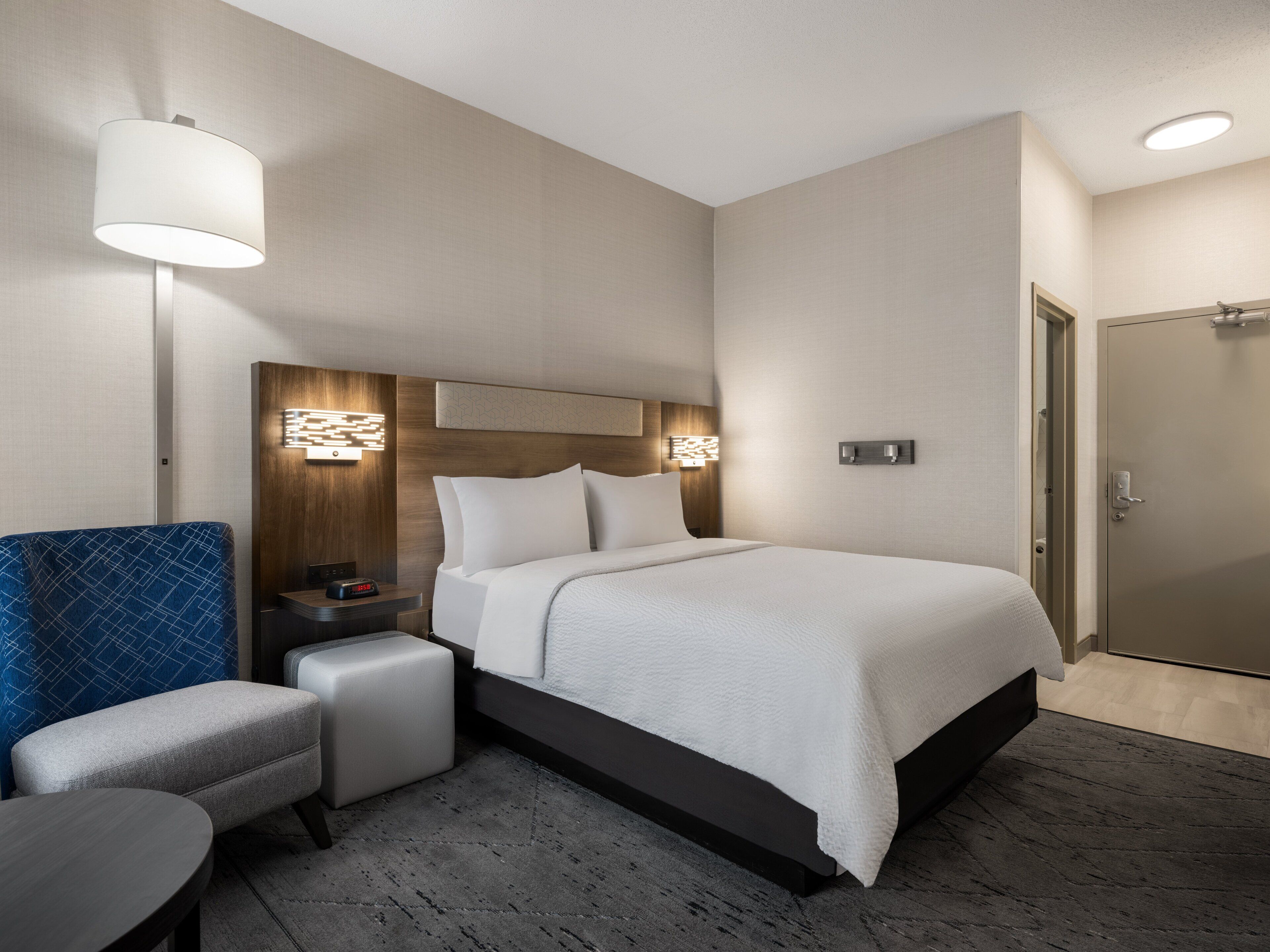 A modern hotel room with a large bed, a bedside table with a lamp, a small seating area, and a door.