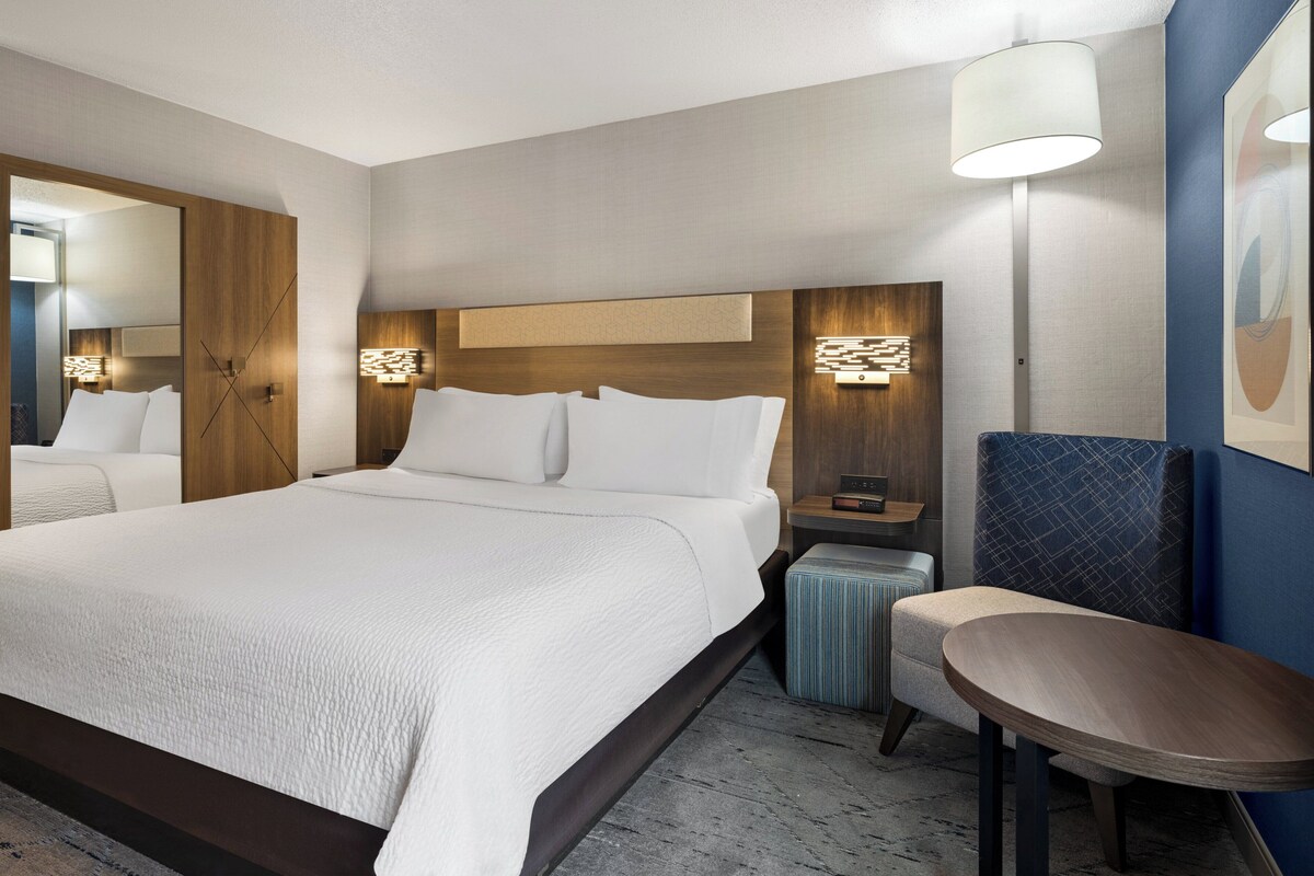 A hotel room with a large bed, a nightstand, a lamp, a chair, and a small table.