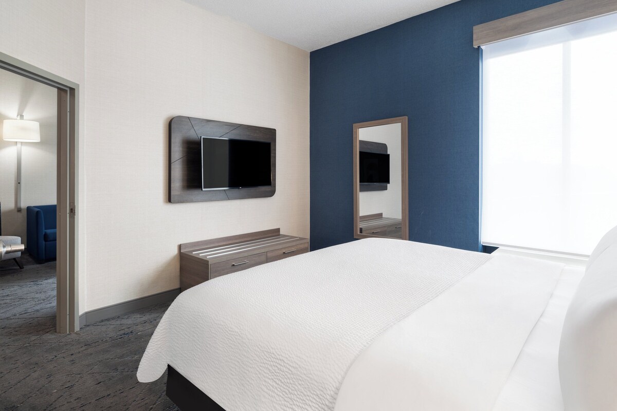 A hotel room with a bed, a television mounted on the wall, a mirror, a bedside table, and a window.