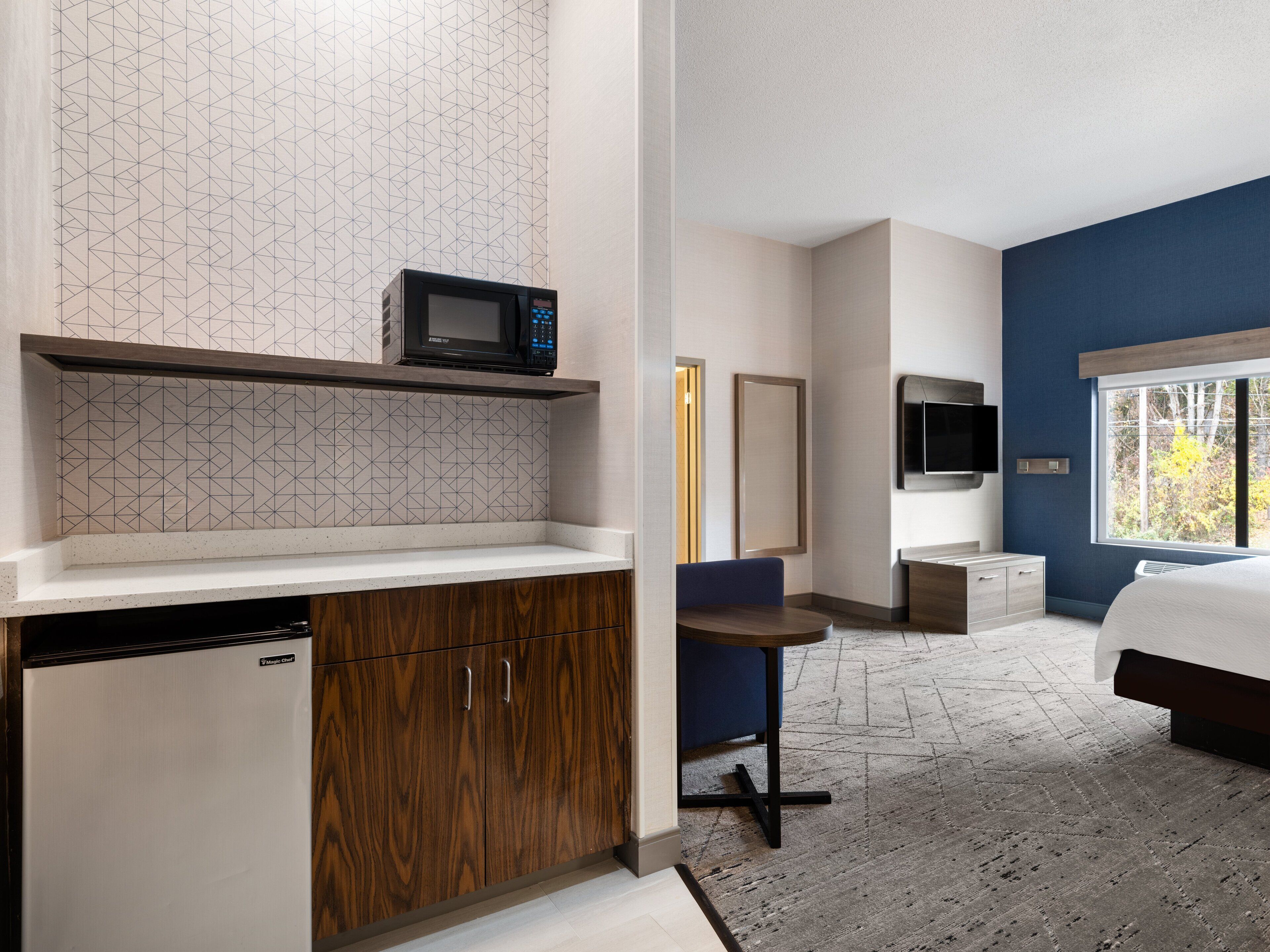 Una habitación de hotel moderna con kitchenette, cama, televisor y ventana con vista a árboles.