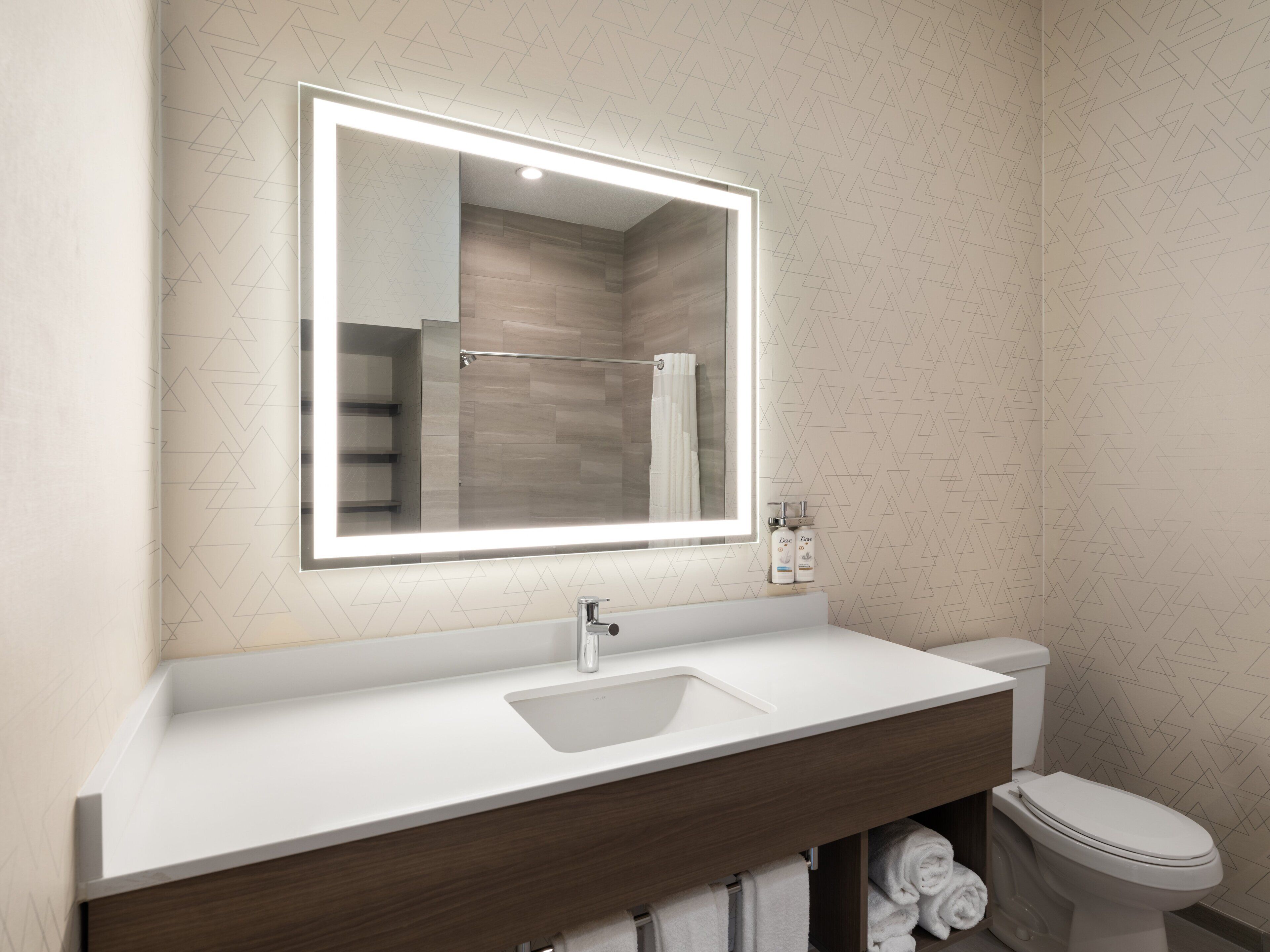 A modern bathroom with a white sink, a mirror with integrated lighting, a wooden vanity, and a toilet.
