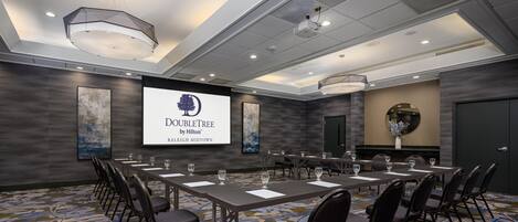 A conference room with a large screen displaying 'DOUBLETREE by Hilton Raleigh Midtown,' a long table with chairs, and a patterned carpet.