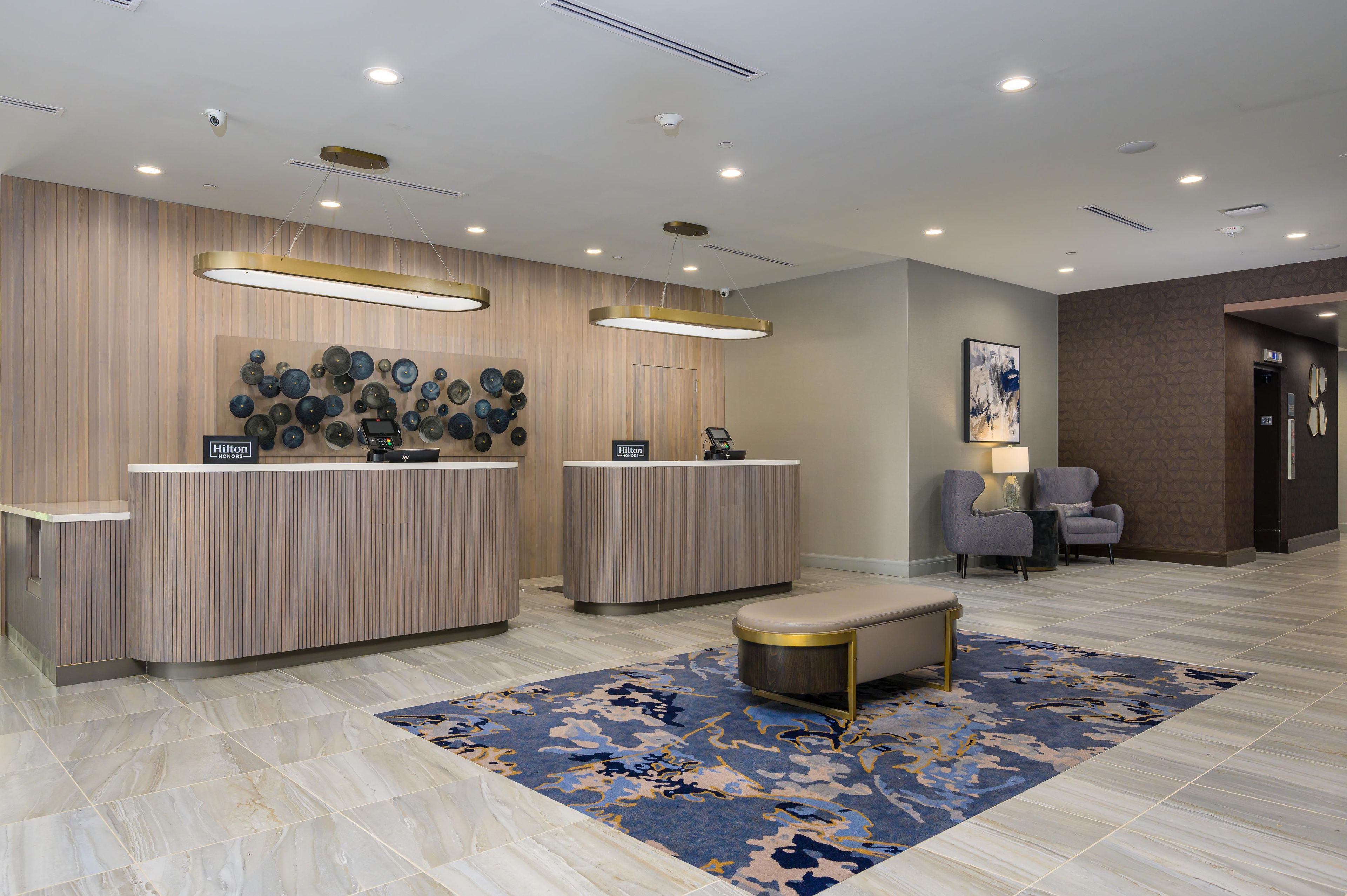 A hotel reception area with two counters, a seating area with chairs, a patterned rug, and a modern ceiling design.