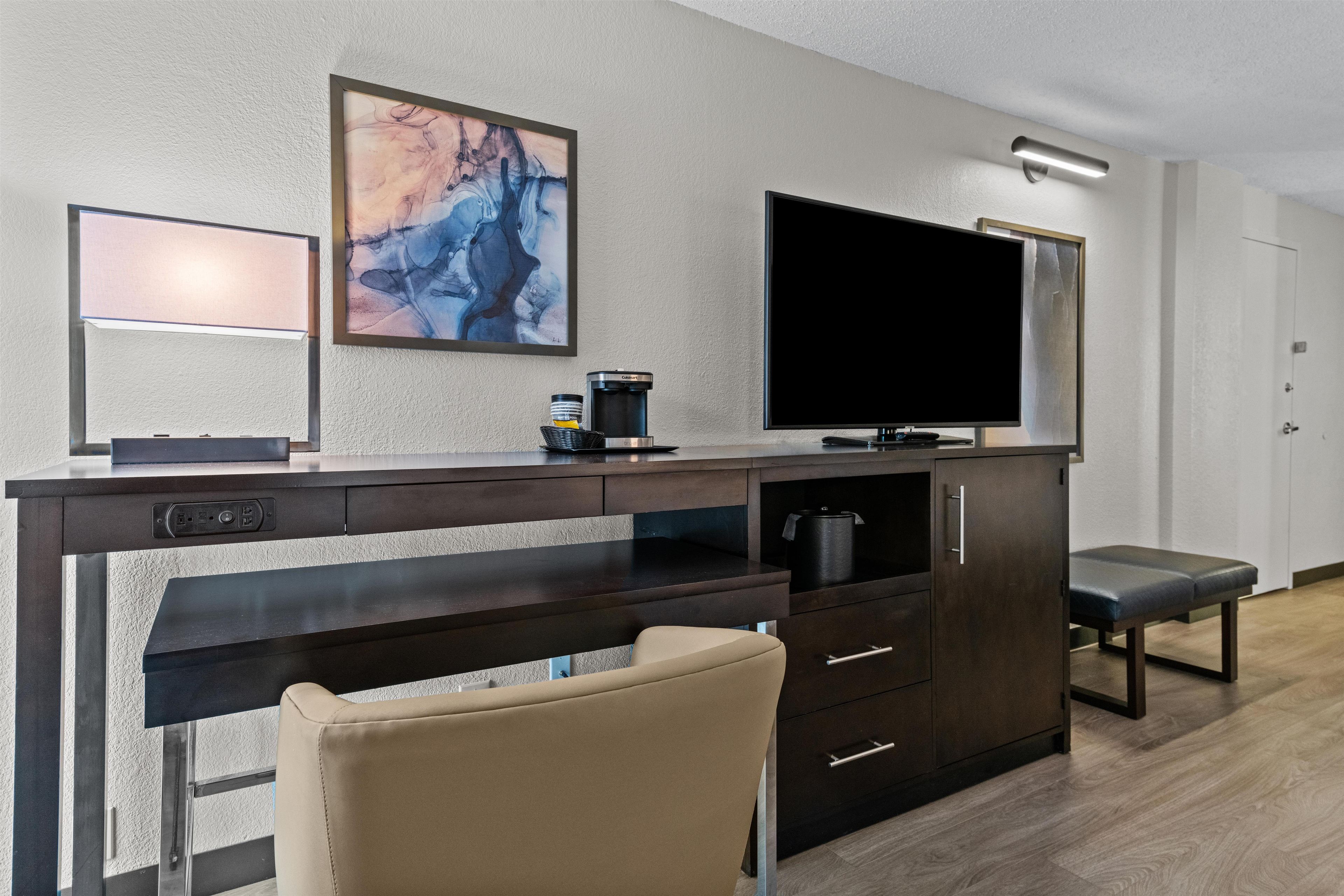 A modern hotel room with a dark wood desk, a flat-screen TV, a coffee maker, and a comfortable chair.