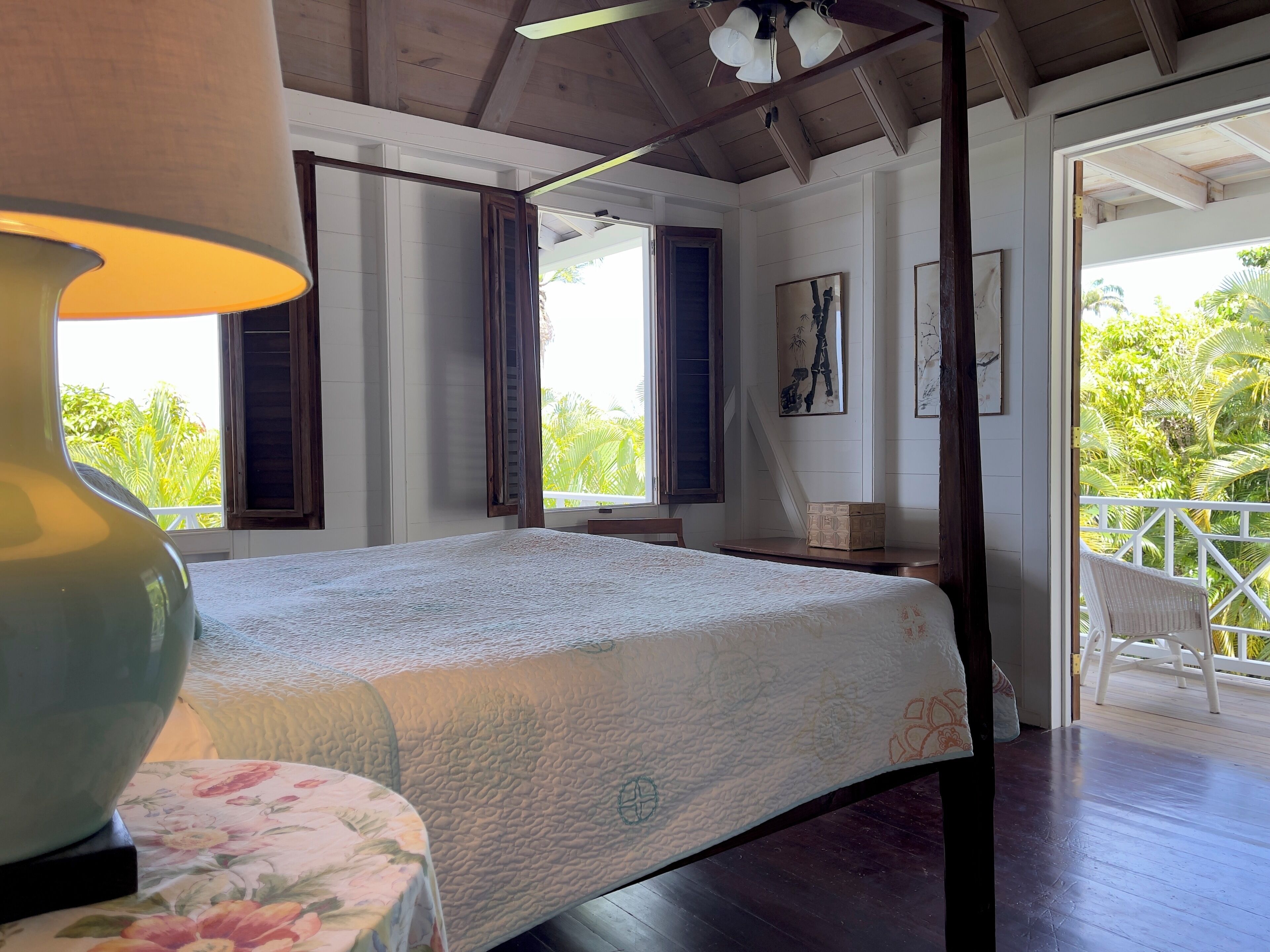 A bedroom with a bed, a table with a floral tablecloth, a lamp, and a balcony with a chair.