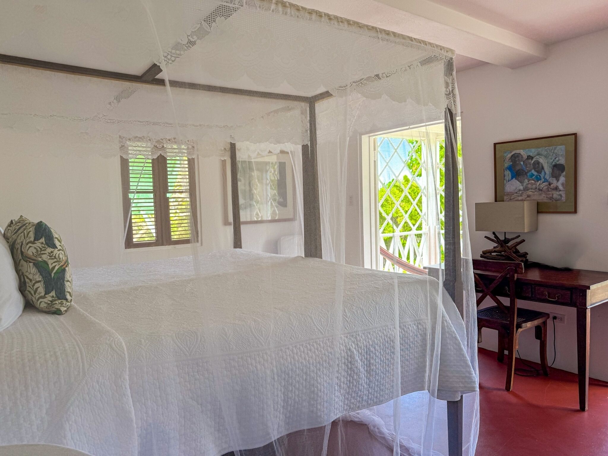 A bedroom with a canopy bed, a desk, and a chair. There are windows with shutters and a painting on the wall.