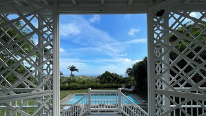 A view through a white lattice structure to a swimming pool with a clear blue sky and palm trees in the background. - Hermitage Nevis (Charlestown)