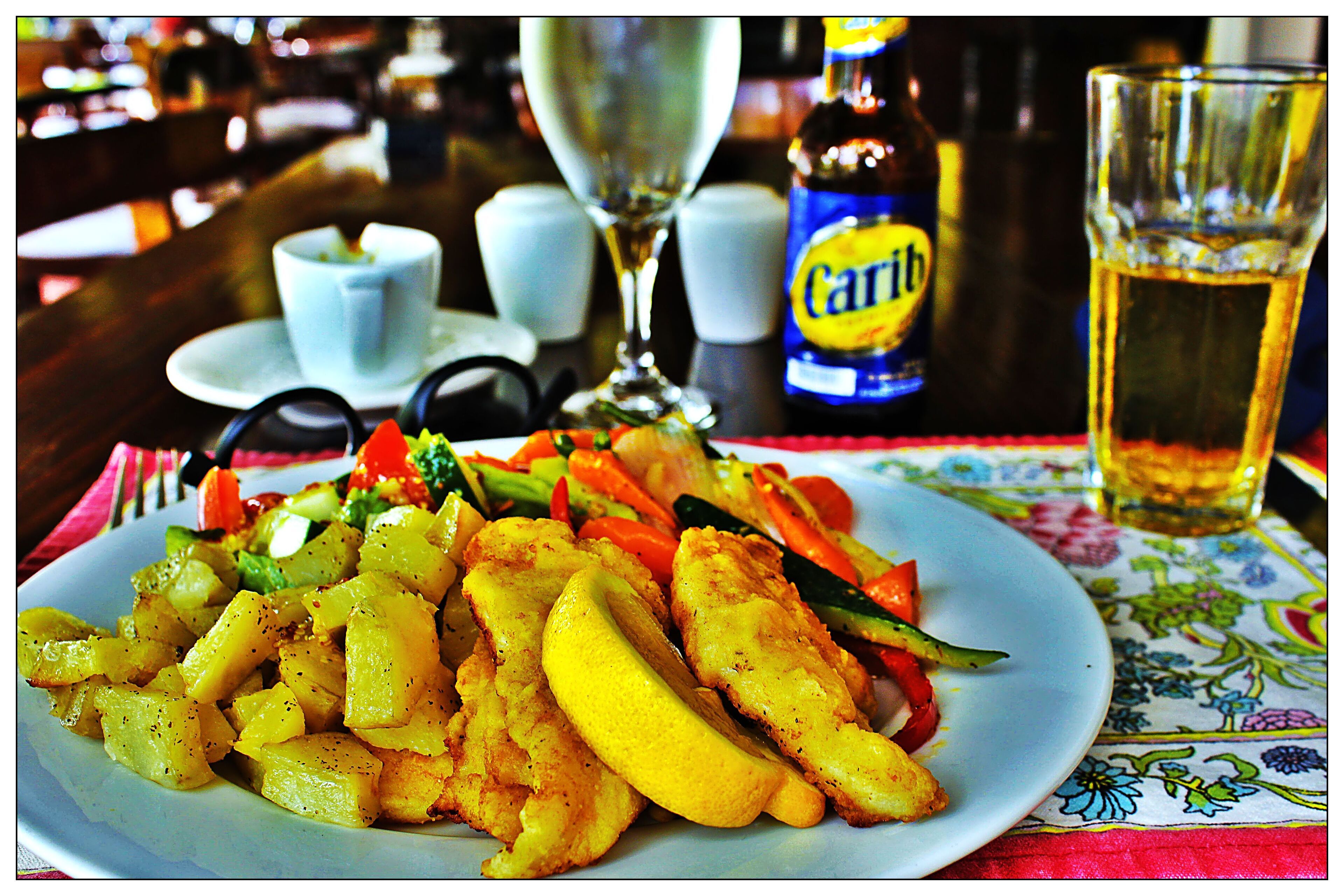 A plate of food with potatoes, carrots, and zucchini, a bottle of Carib beer, a glass of beer, and a wine glass on a table.