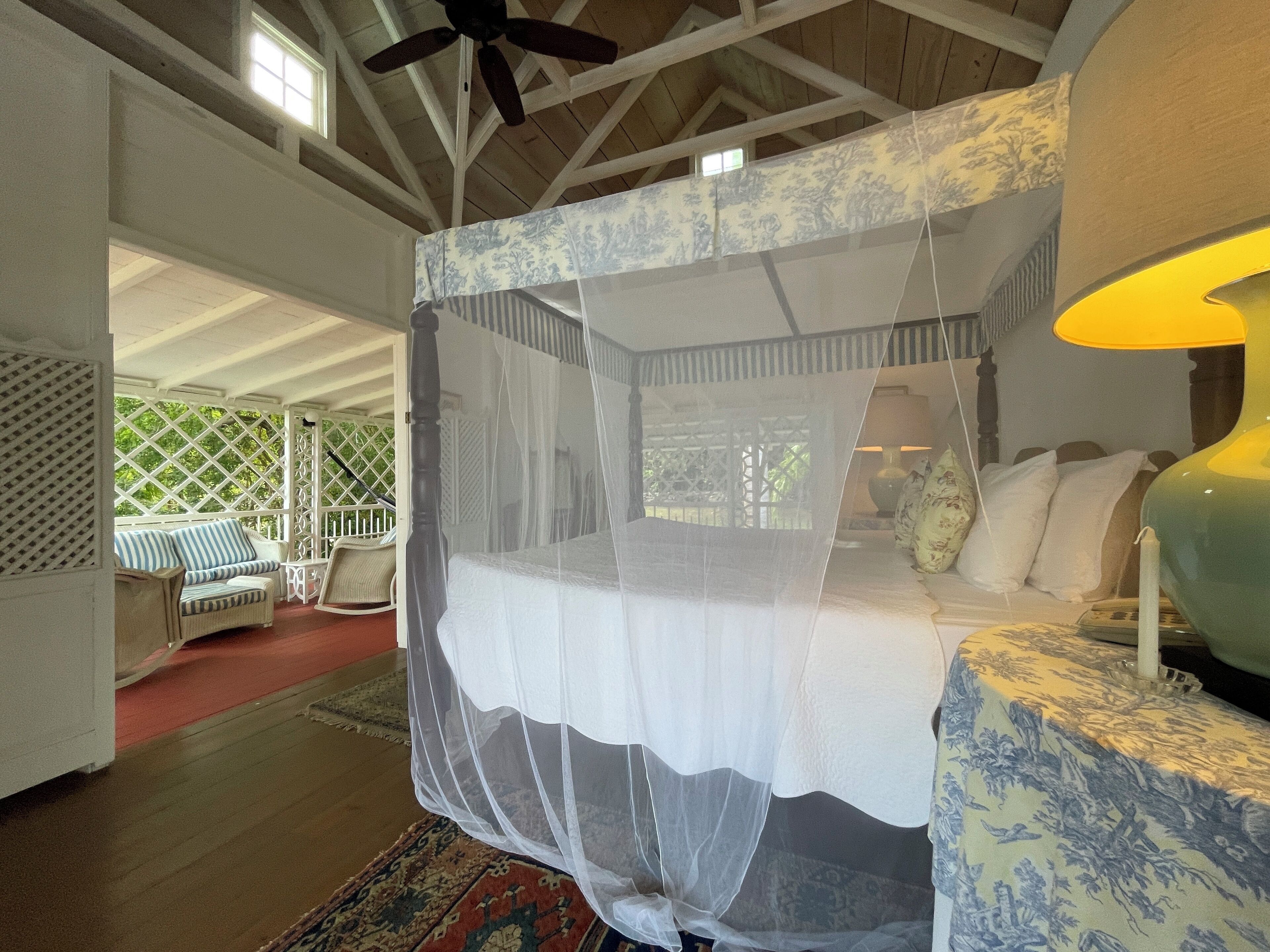 A bedroom with a four-poster bed, a canopy, a nightstand, a lamp, and a view of the outdoors.